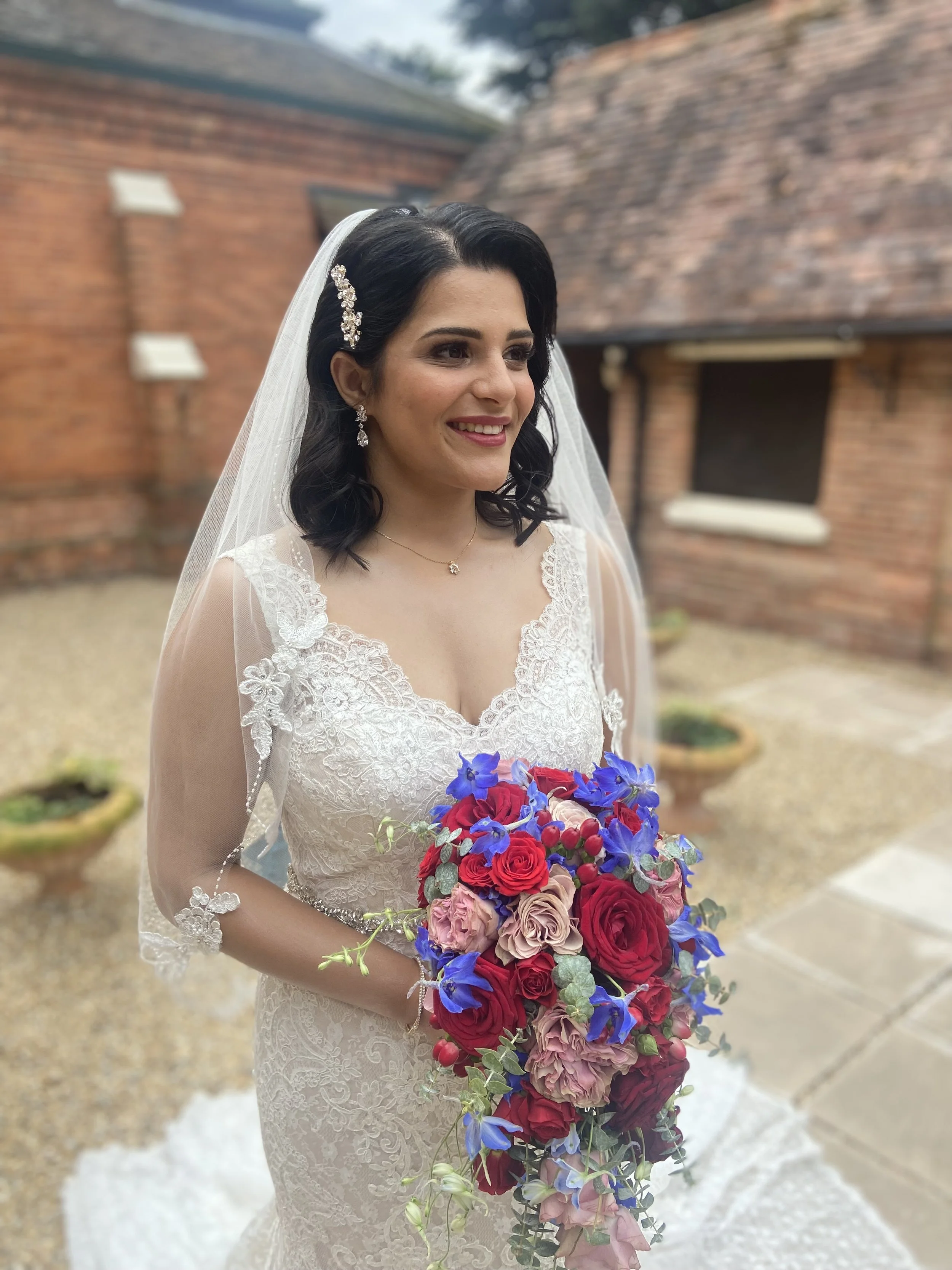 A bride in a lace wedding dress holding a colorful bouquet, standing outdoors near a brick building.