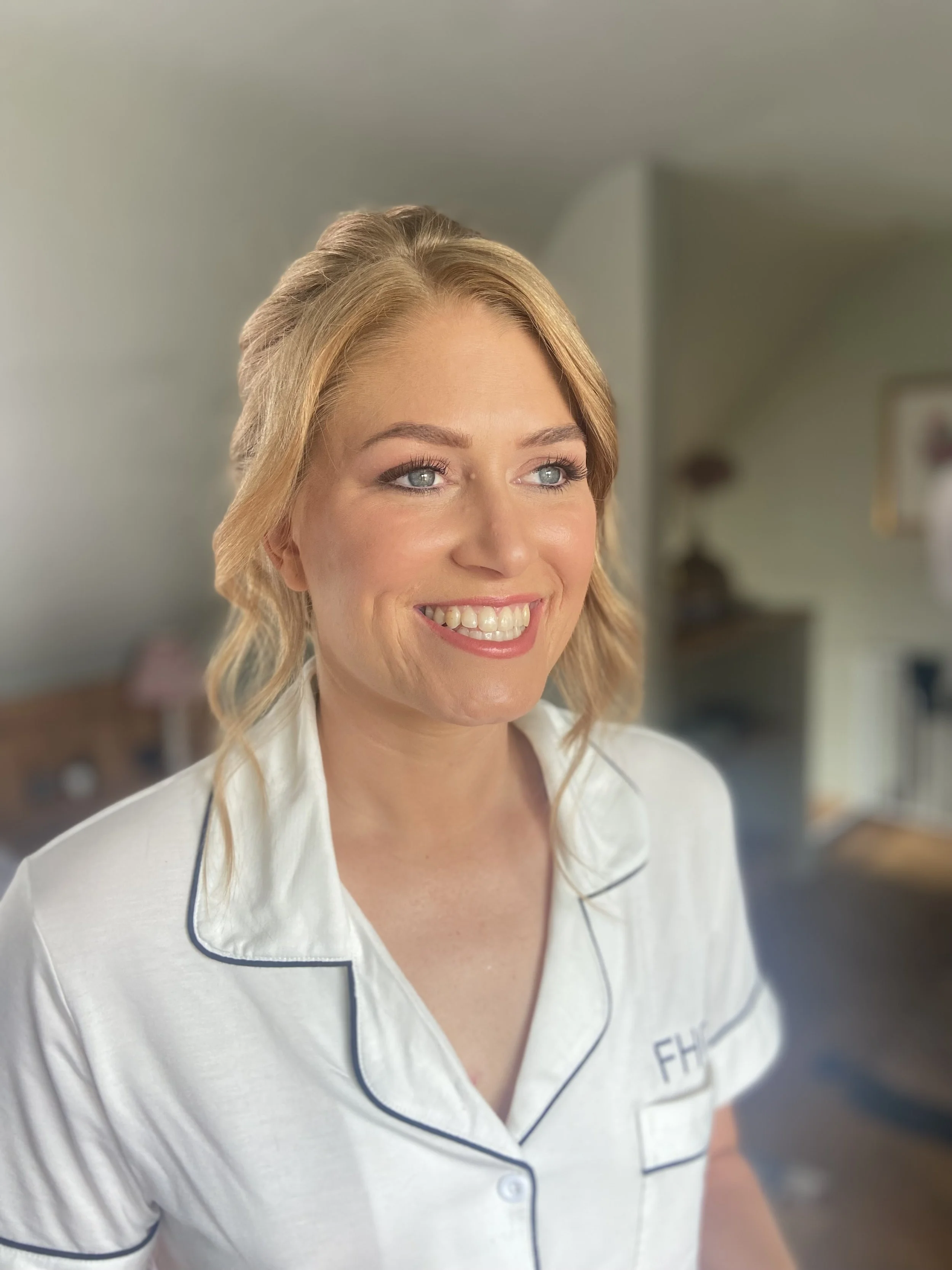 A smiling woman with blonde hair wearing white pajamas with navy piping, standing in a bright living room.