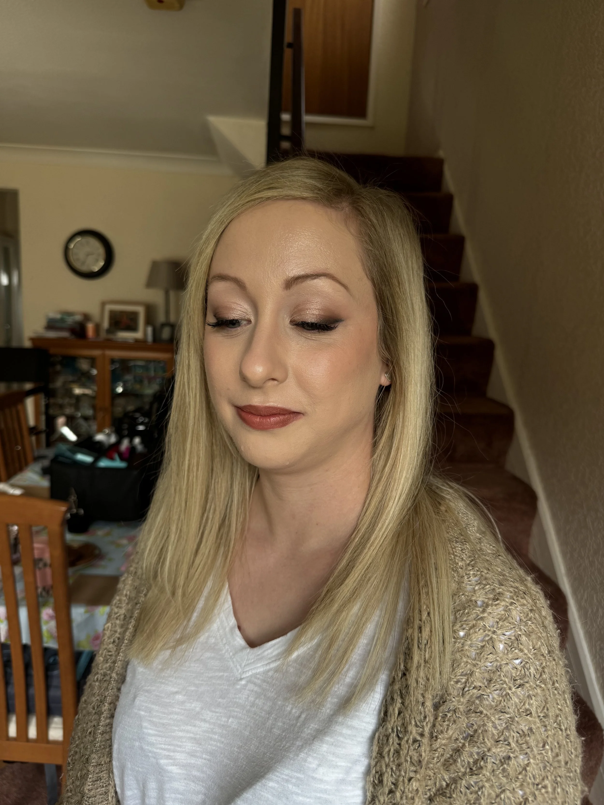 A woman with blonde hair wearing makeup and a beige knitted cardigan standing in a home interior with stairs in the background.