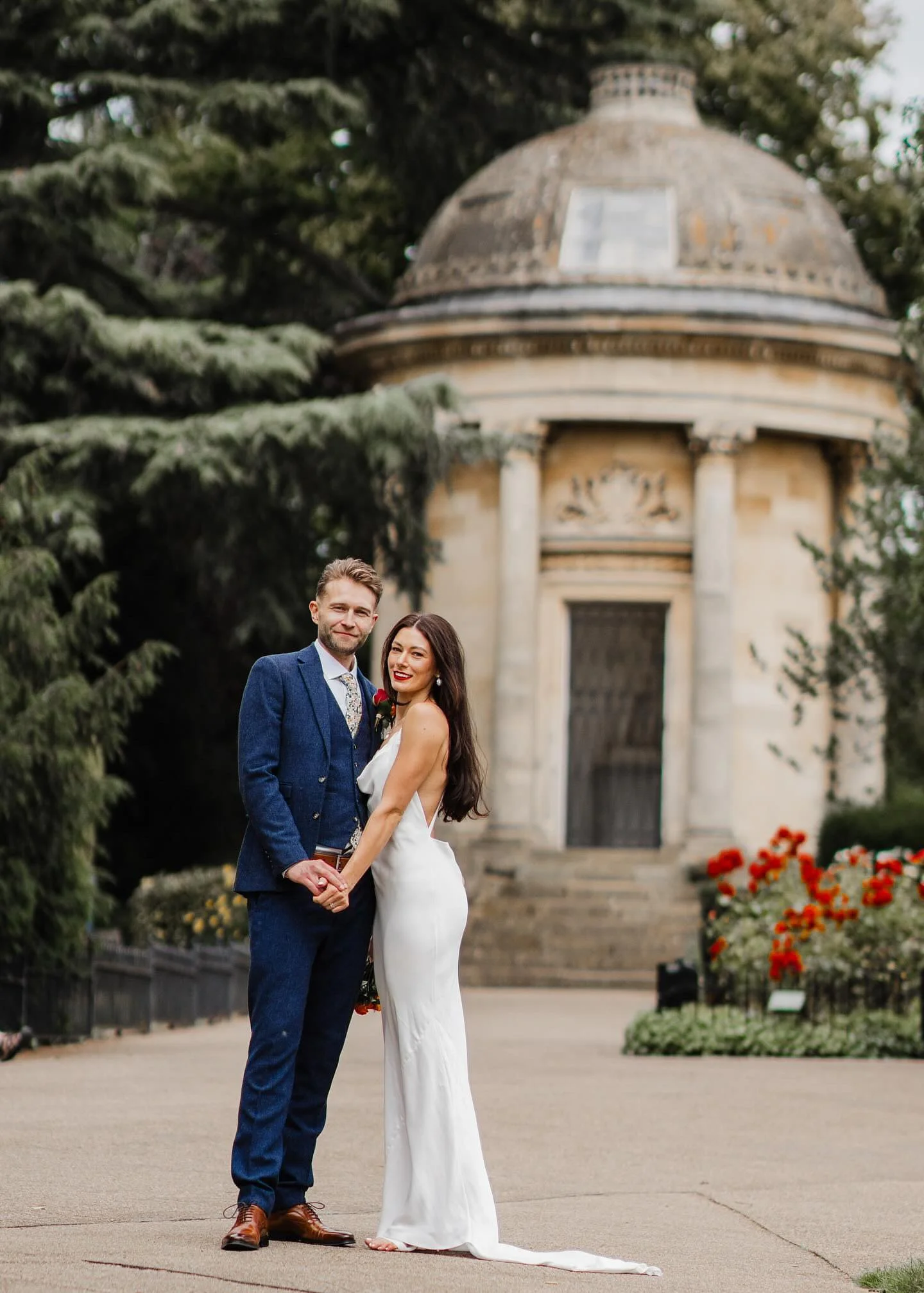 A newlywed couple holding hands and smiling at the camera in front of a historic round stone pavilion with a domed roof, surrounded by trees and flower beds.