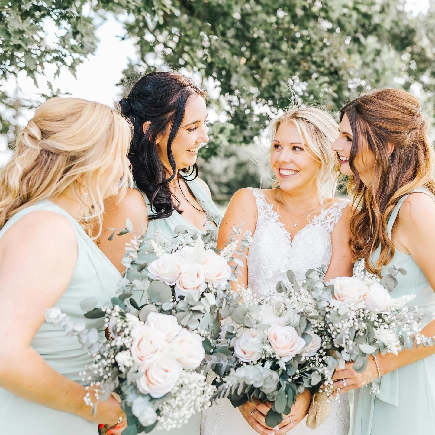 A bride with blonde hair in a white lace wedding dress holding a bouquet of white and pink roses and greenery, surrounded by four women in pastel green dresses, outdoors with trees in the background.