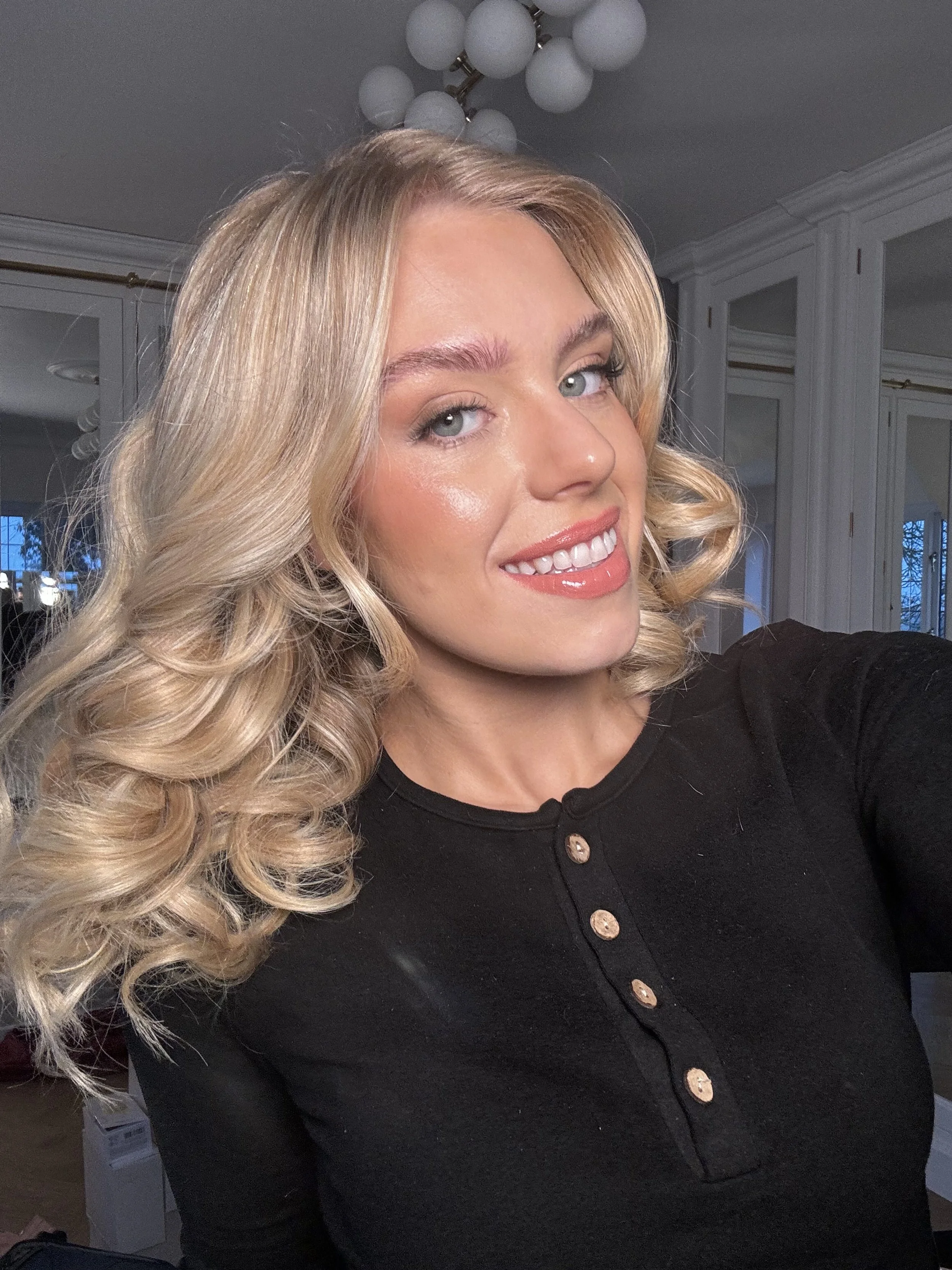 A young woman with blonde, curly hair taking a selfie indoors. She is smiling and wearing a black top with gold buttons. Behind her is a modern room with large windows, white walls, and a decorative white spherical ceiling light.