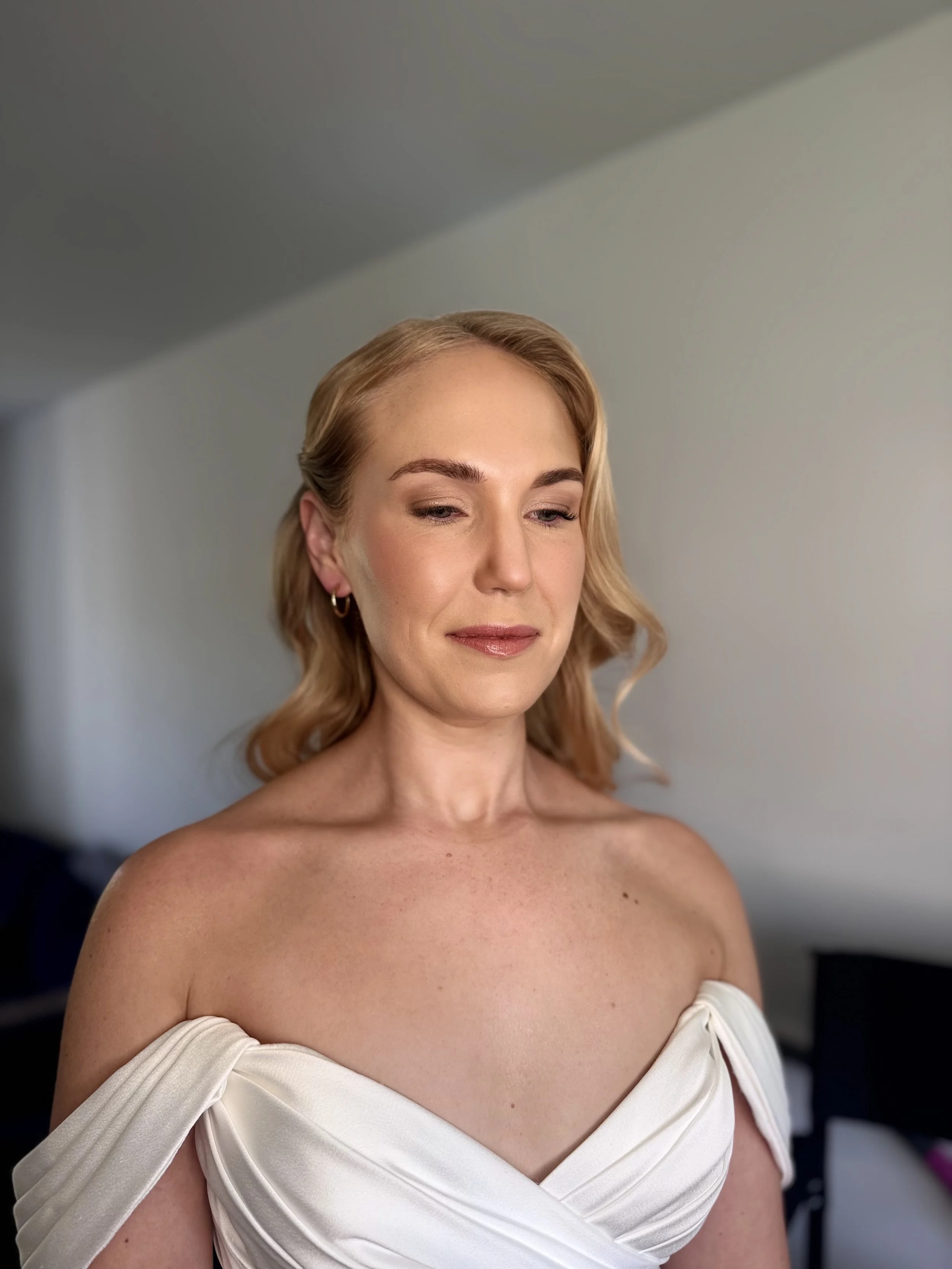 A woman with blonde hair and light makeup wearing an off-shoulder white dress, standing indoors with a neutral background.