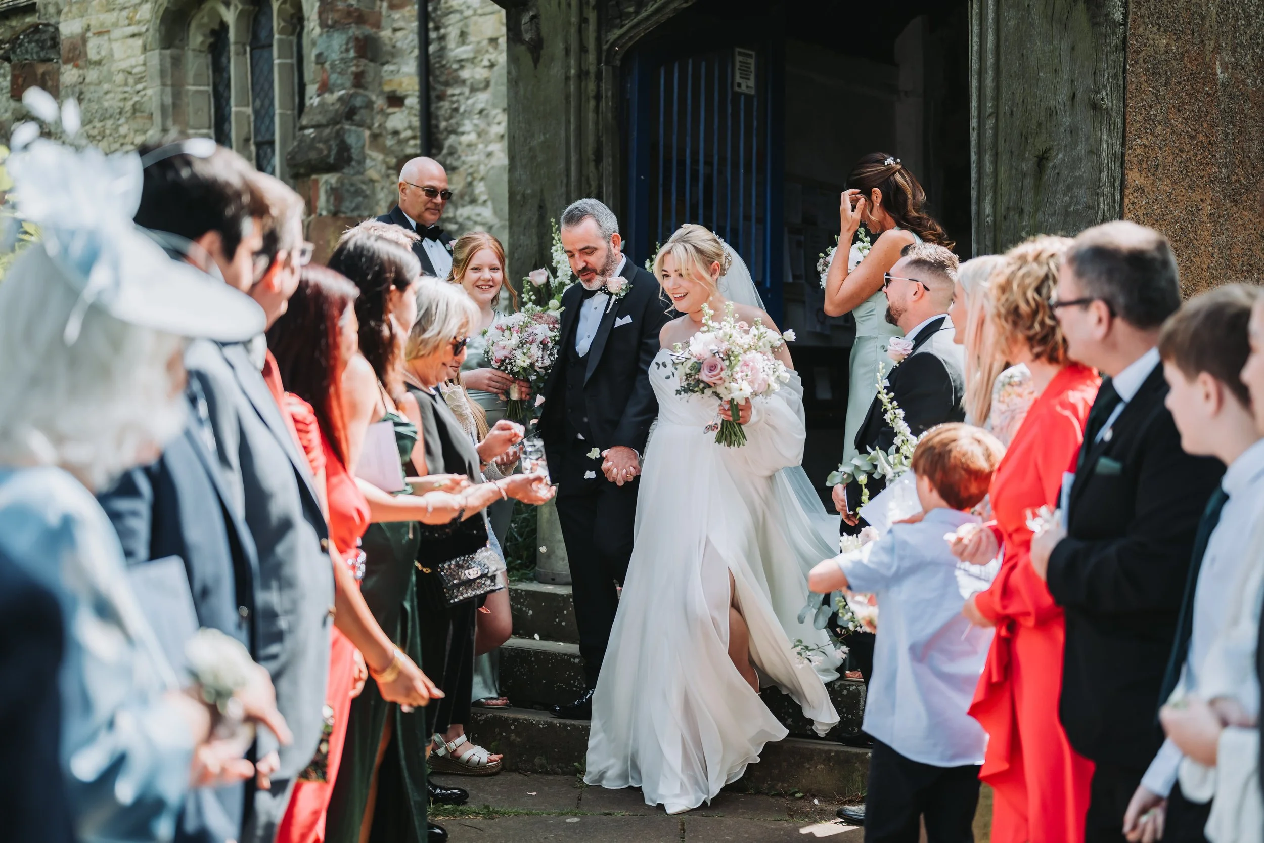 Bride and groom holding hands descending stairs outside surrounded by guests throwing flower petals.