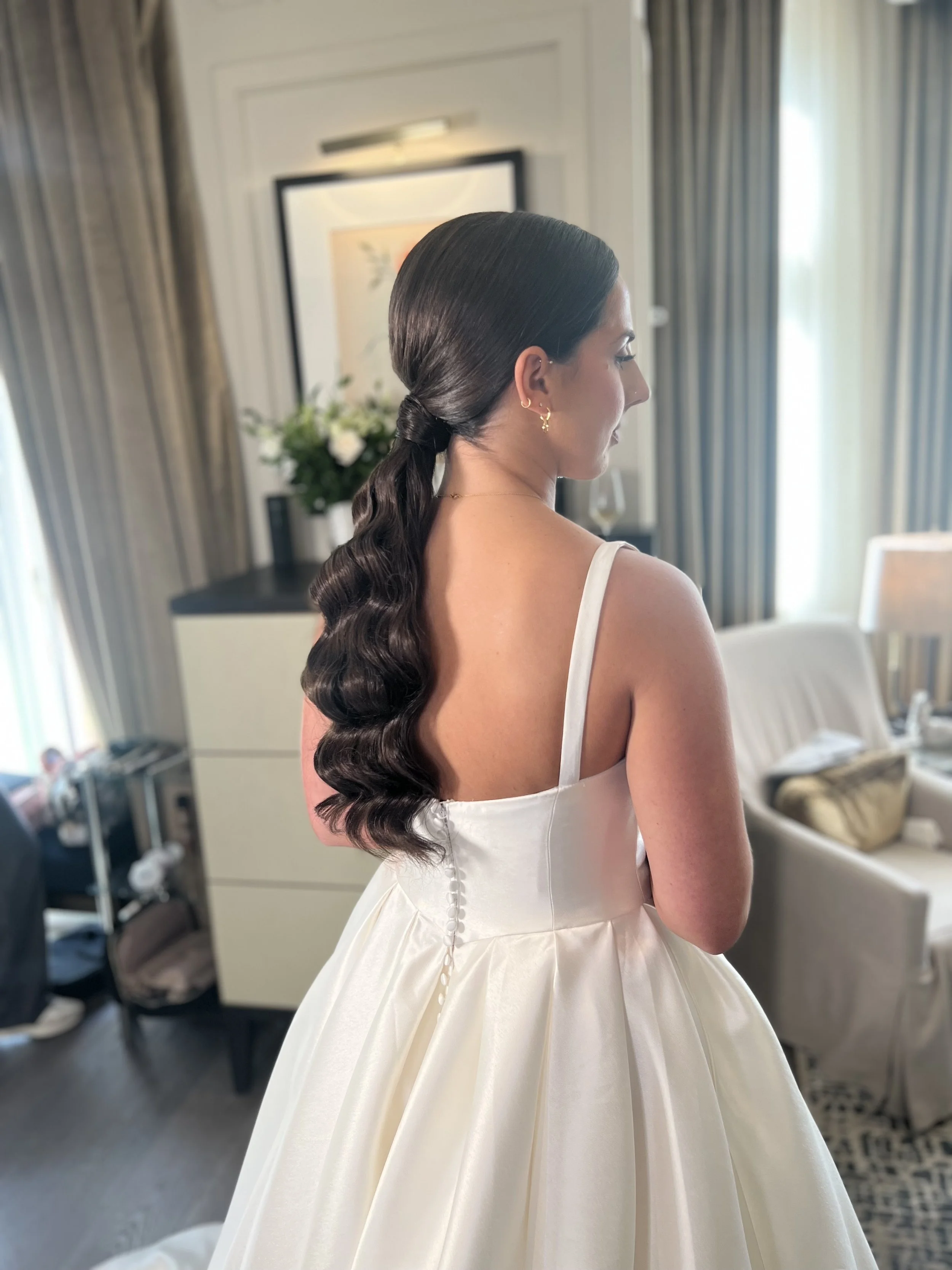 Woman in a white wedding dress with a gathered full skirt, standing in a well-lit room with curtains and furniture, seen from the side.