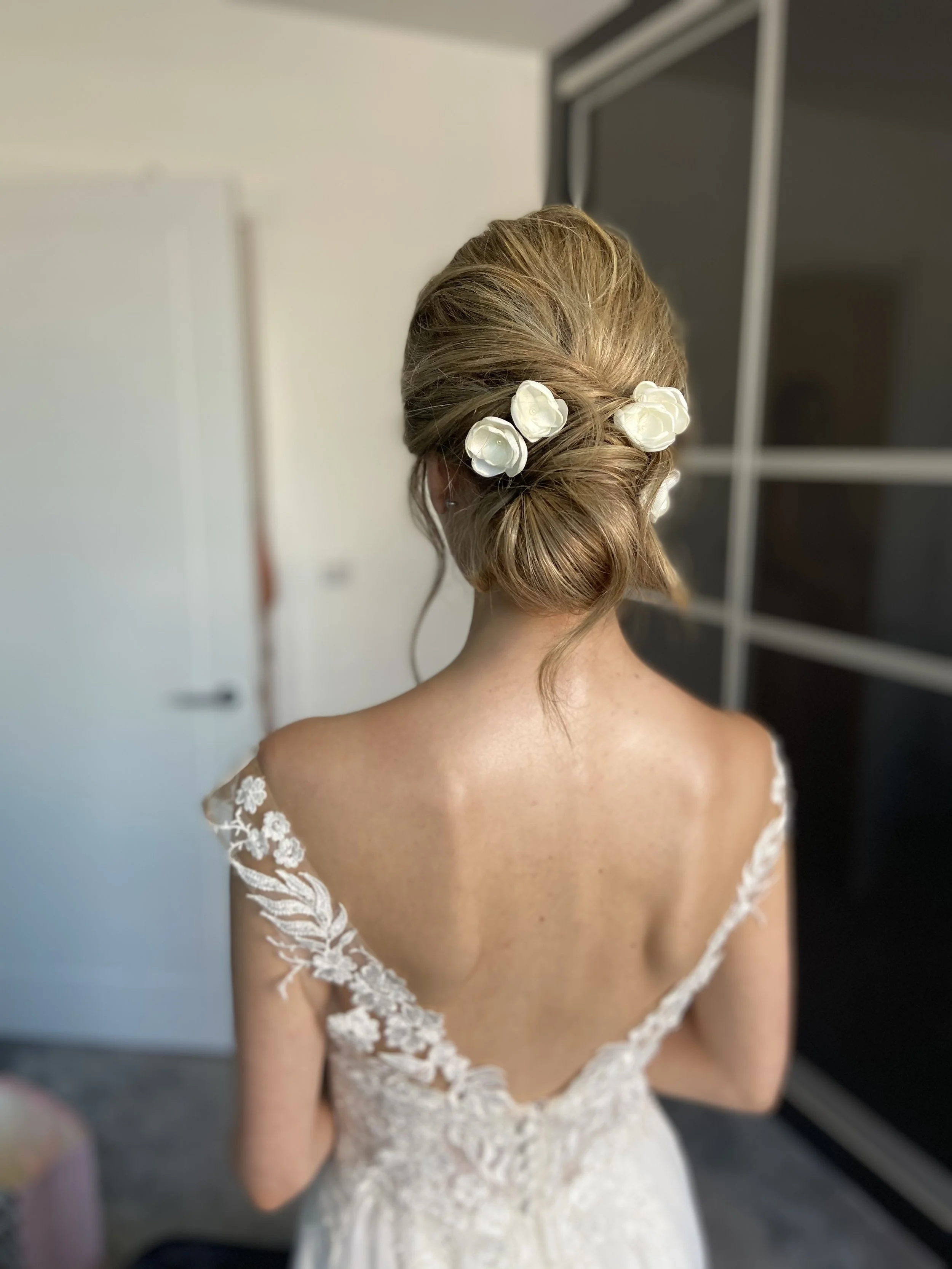 Back view of a bride with styled hair adorned with white flowers, wearing a lace wedding dress with floral embroidery on the shoulders.