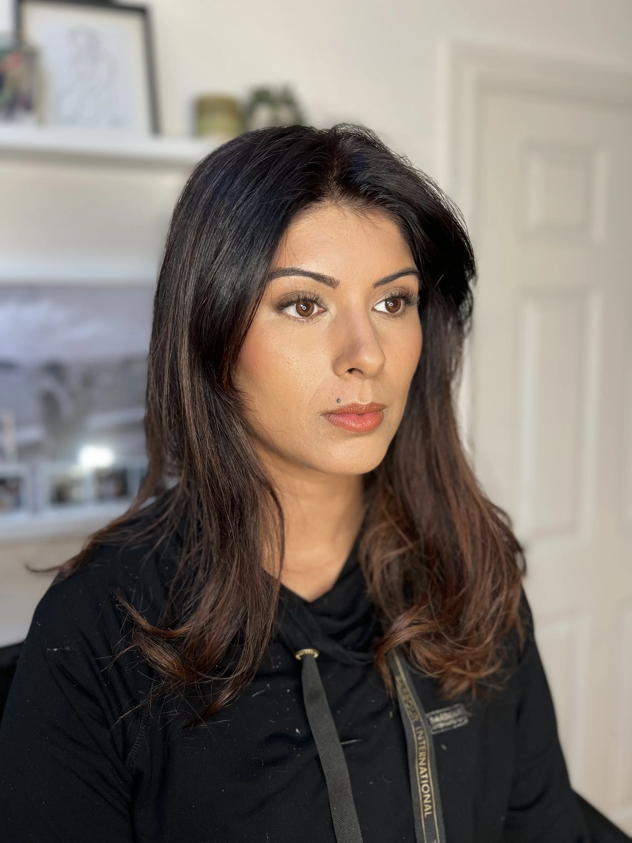 A woman with long dark hair featuring brown highlights, wearing a black top with a gold zipper and a black lanyard, sitting indoors in front of a blurred background with shelves and framed photos.