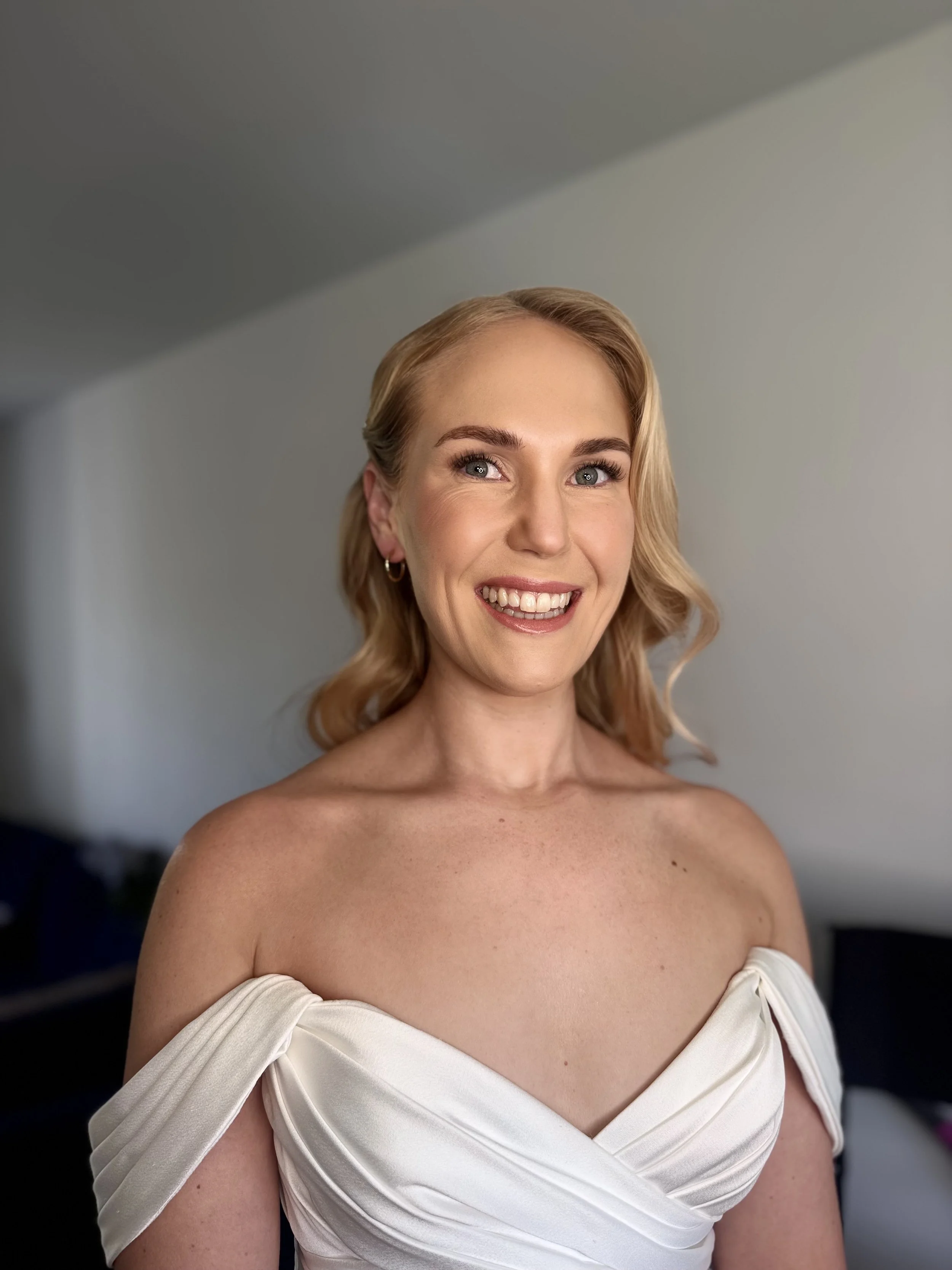 A smiling Bride with blonde hair and blue eyes wearing a white off-the-shoulder dress, in a room with a plain light-colored wall.