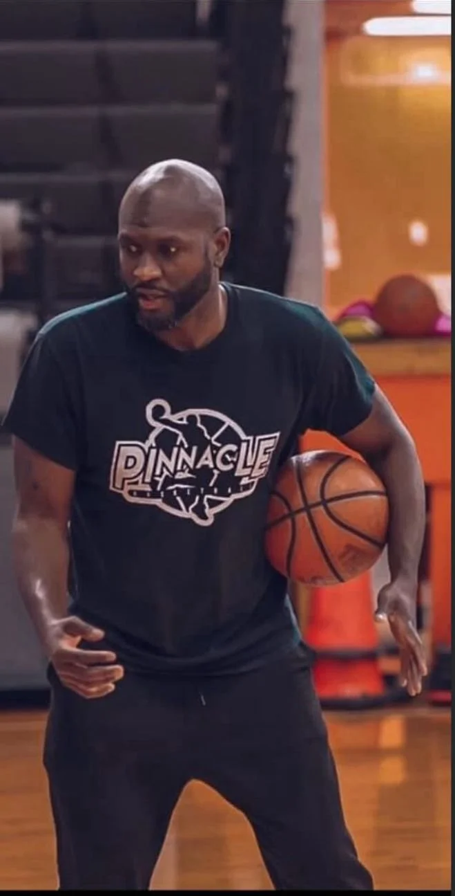 A man in a black Pinnacle basketball shirt is holding a basketball while on a basketball court.