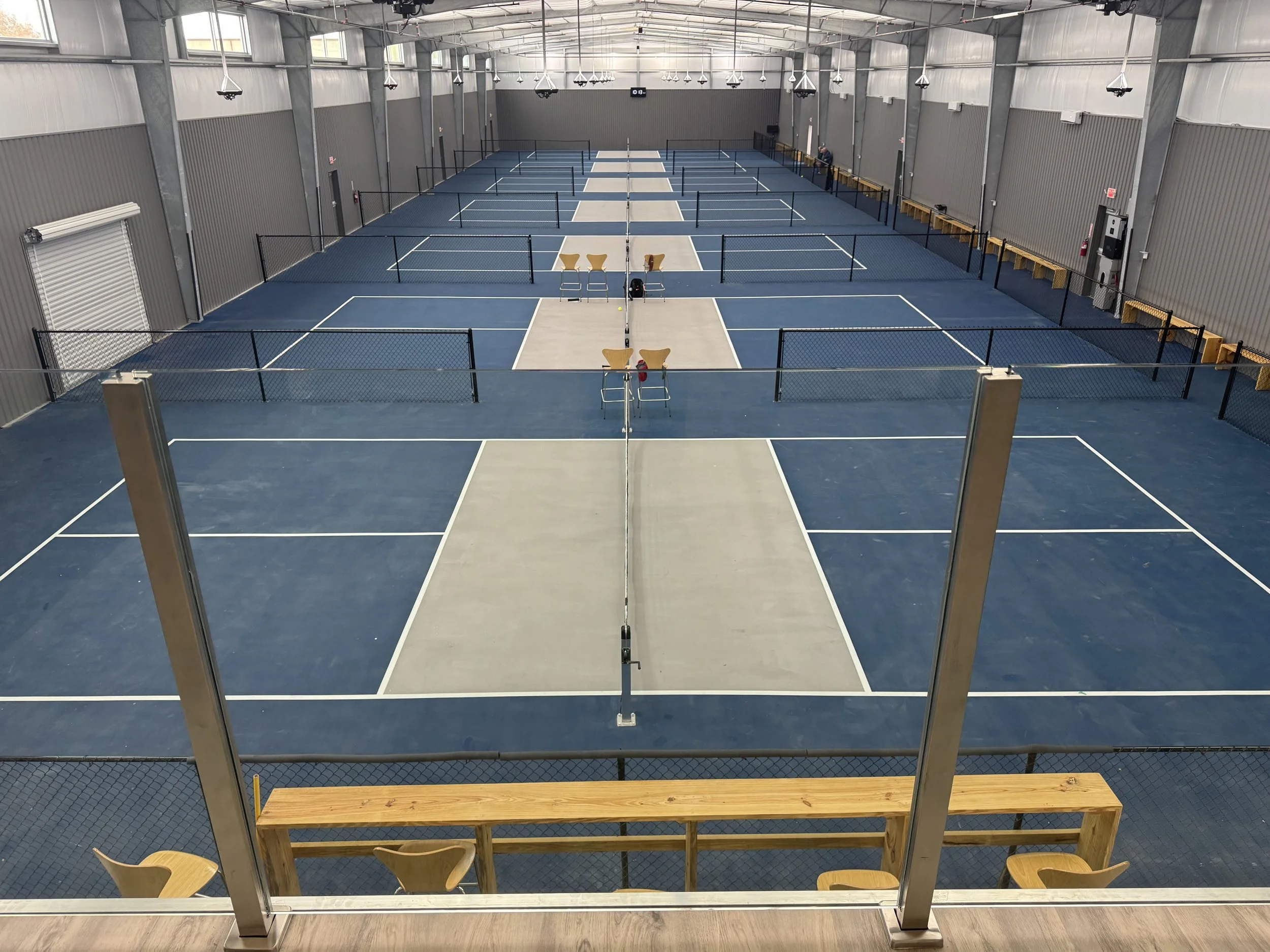 Multiple indoor tennis courts with blue surfaces, separated by black nets, in a large sports facility with high ceilings and gray walls. There are empty courts, a few chairs and benches along the sides, and bright lighting.