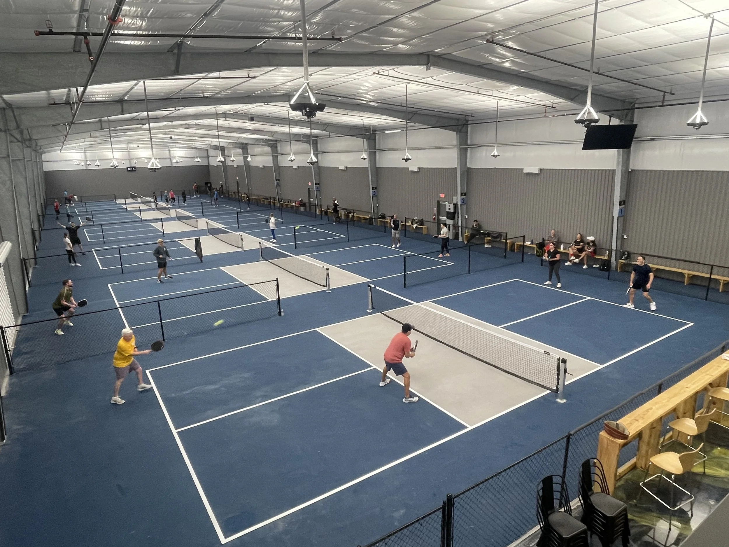 Indoor pickleball court with multiple games in progress and players of various ages playing and sitting along the sides.