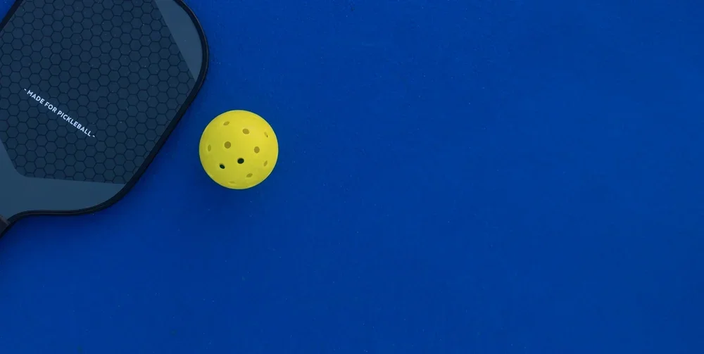 A yellow pickleball with holes on a blue pickleball court, with a pickleball paddle partially visible.