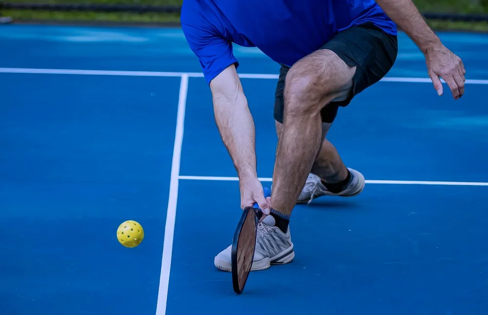 Man in athletic clothing playing pickleball on an outdoor blue court, reaching to hit a yellow perforated ball with a paddle.