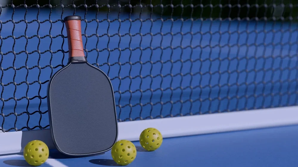 A tennis paddle leaning against a net on a tennis court, with three yellow tennis balls in front.