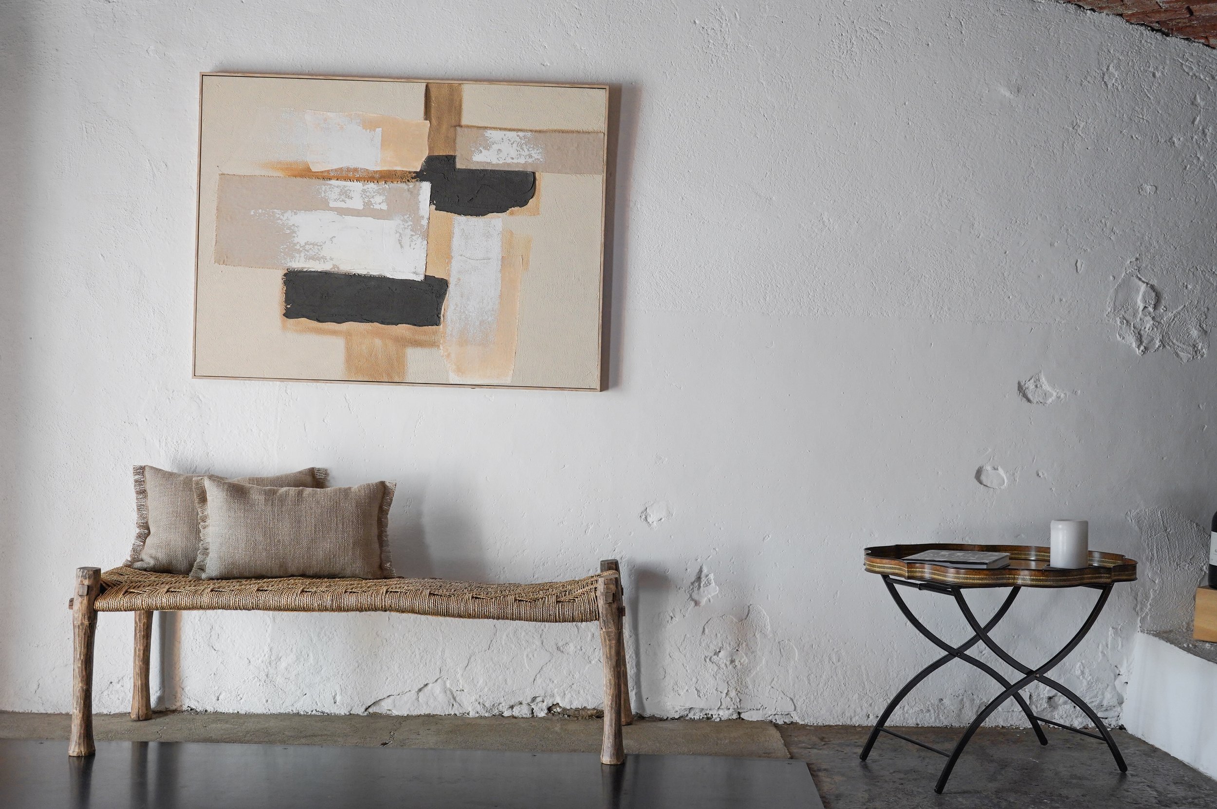 Minimalist room with abstract art, woven bench with cushions, and a side table with a candle.