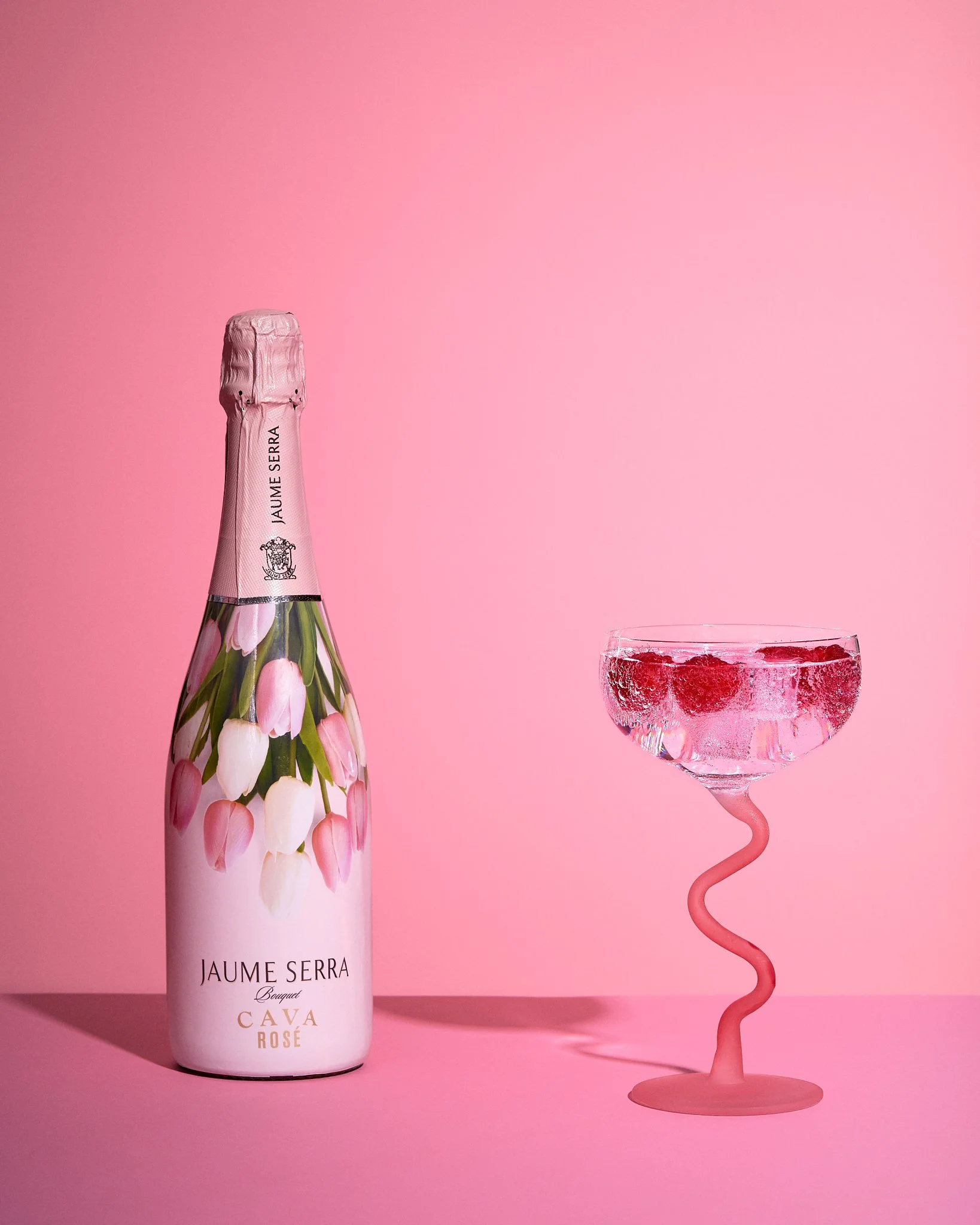 Vibrant colourful image of a bottle of pink Cava next to a pink cocktail glass with a bendy stem containing fizz and raspberries against a pink background. Hard shadows.