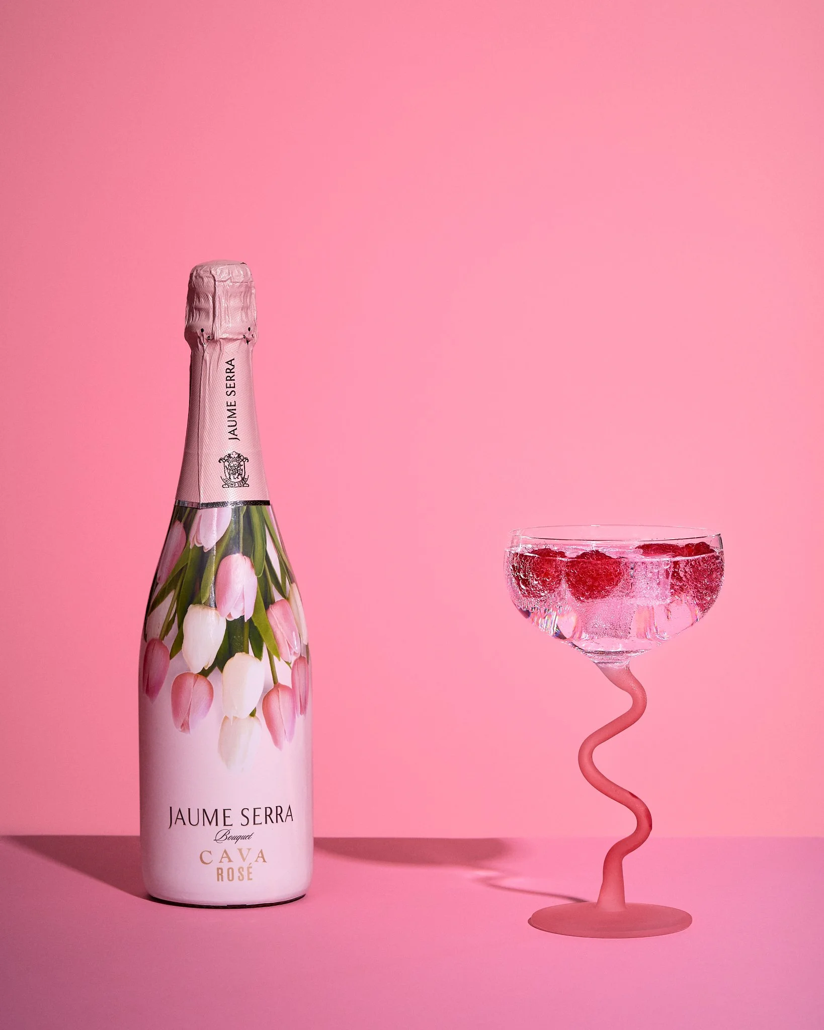 Vibrant colourful image of a bottle of pink Cava next to a pink cocktail glass with a bendy stem containing fizz and raspberries against a pink background. Hard shadows.