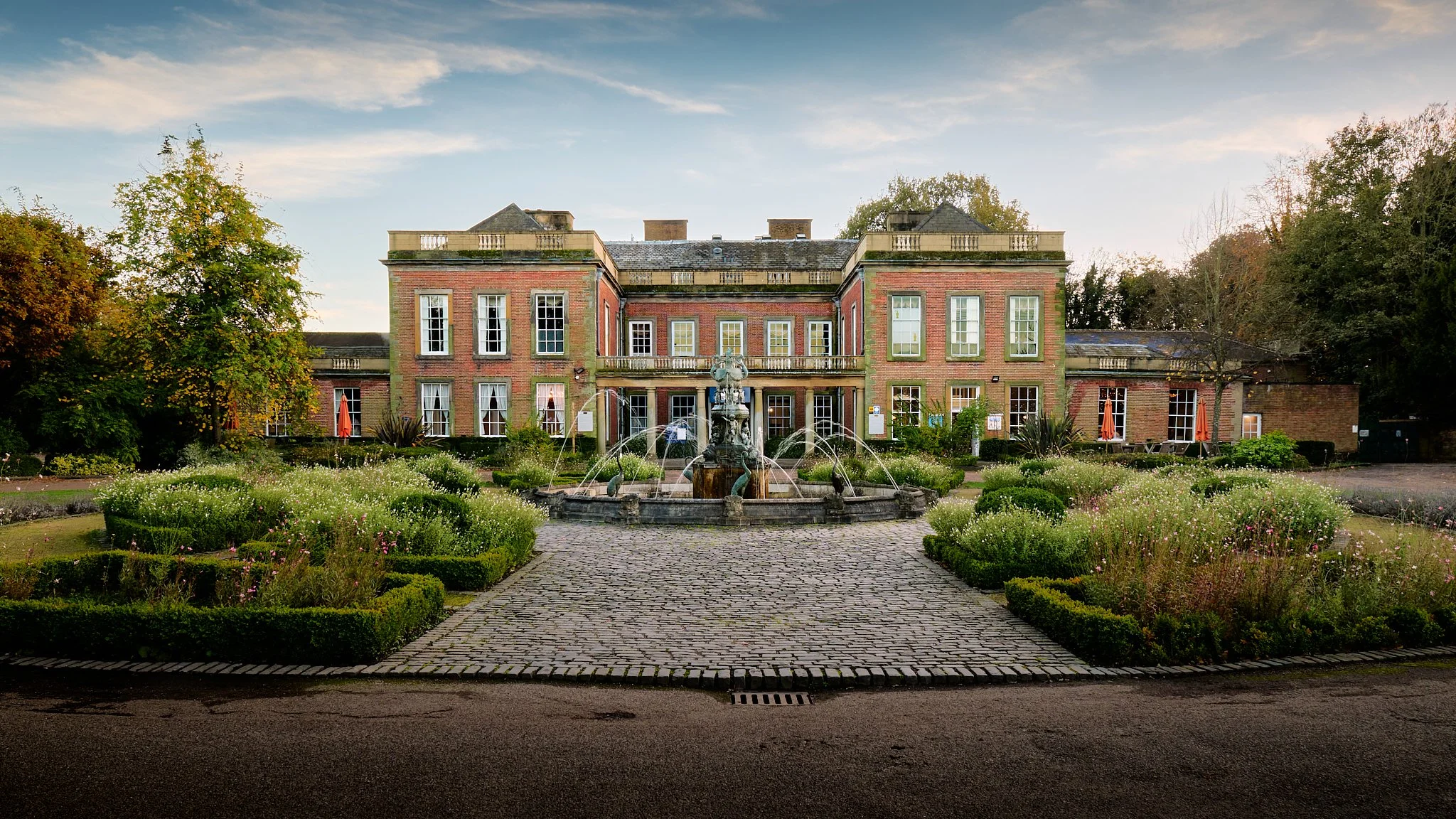 A large red-brick mansion with tall, white-framed windows, surrounded by a well-maintained garden with a fountain in the center, and trees on either side. The sky is partly cloudy with sunlight.