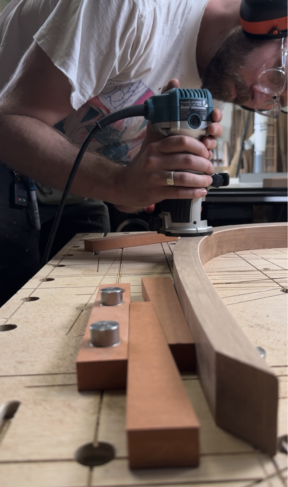 Close-up of a woodworker routing the edge of a curved wooden component secured to a workbench.