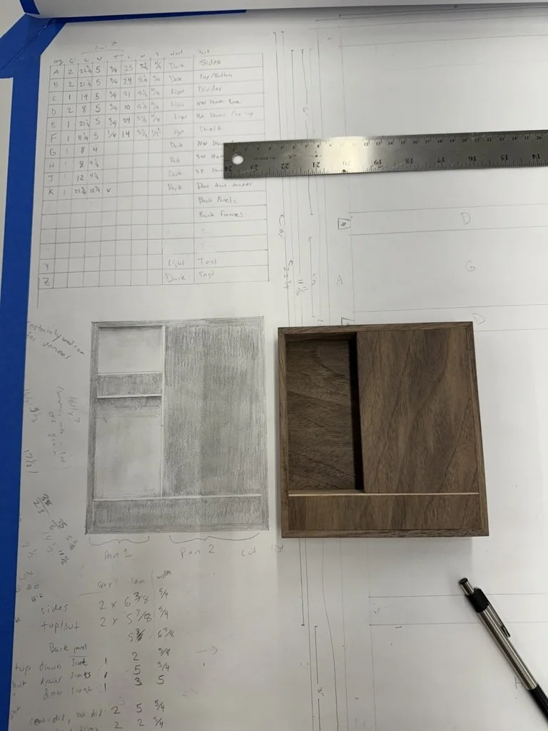 Drafting paper with hand-drawn woodworking plans, measurements, and a ruler, next to a small wooden cabinet model.