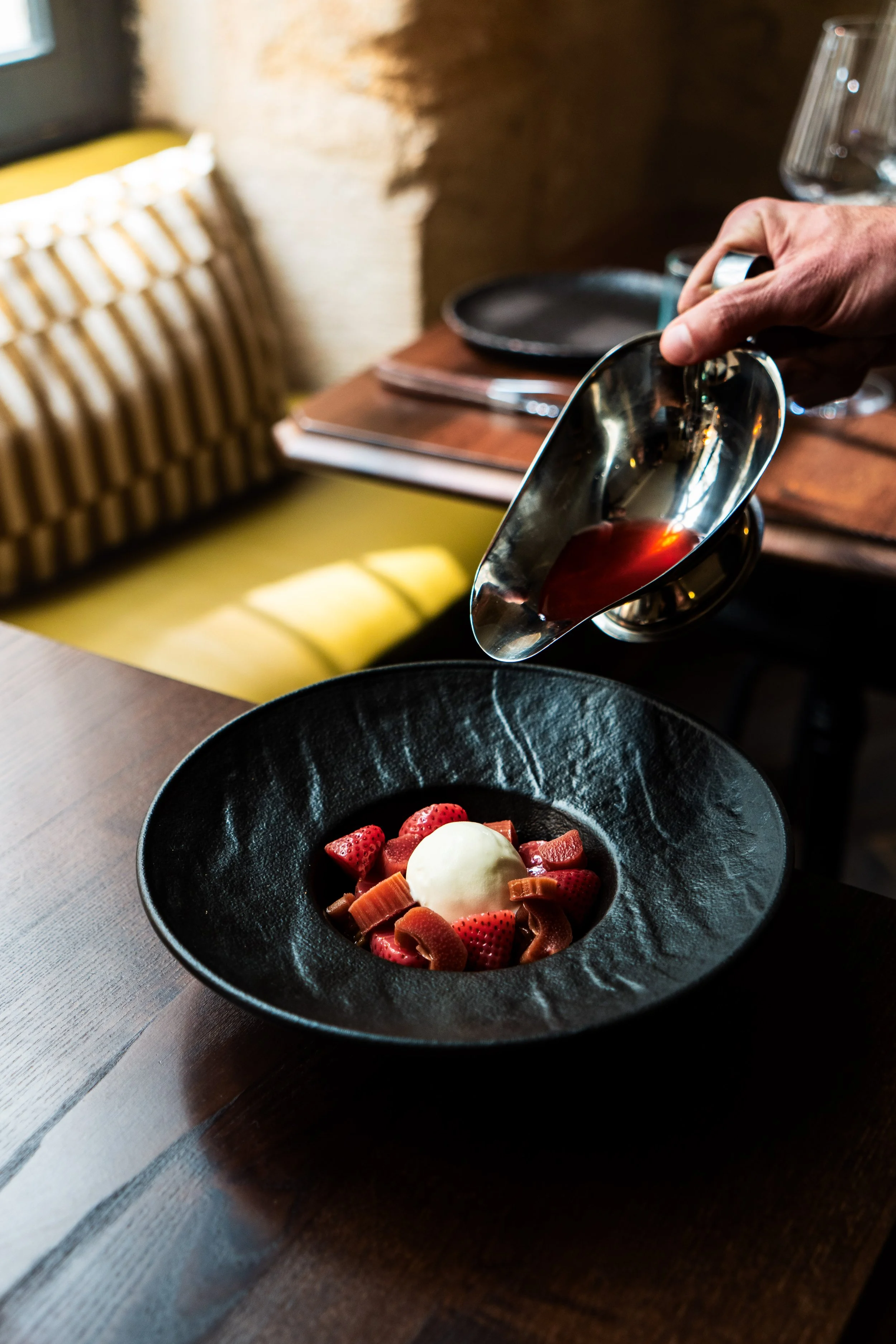 Une main verse du sirop rouge sur un dessert de fraises, crème glacée et rubans de caramel dans un bol noir.