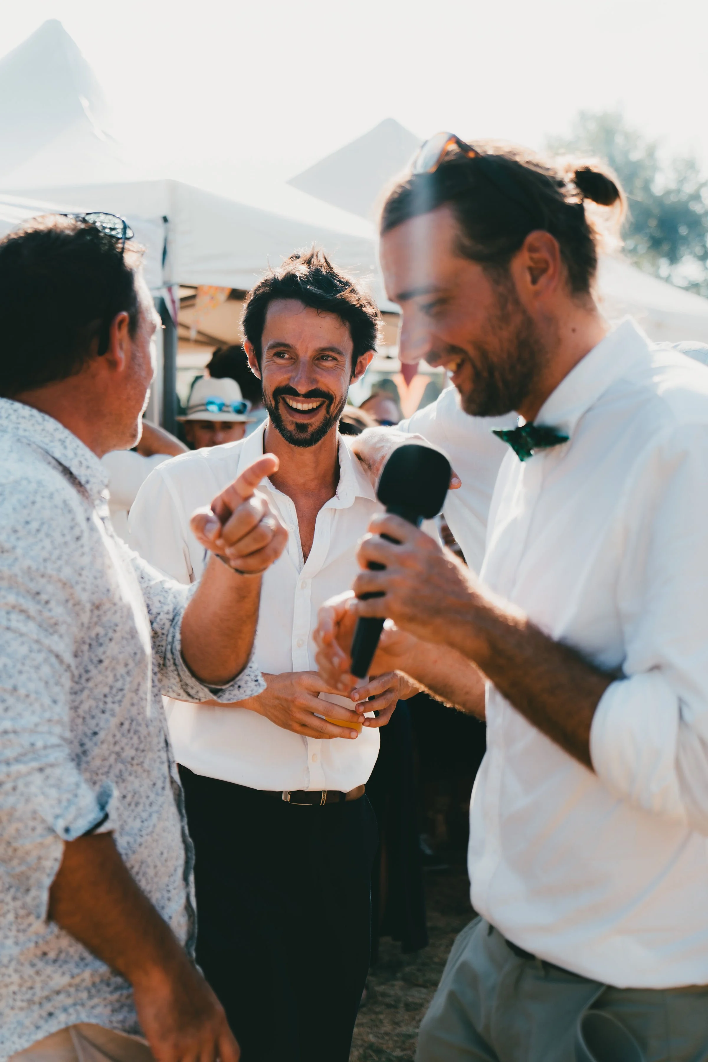 Trois hommes, un en costume blanc avec un nœud papillon, un autre avec une chemise à motifs et un troisième avec une chemise blanche, discutent et rient lors d'un événement en extérieur, avec des tentes blanches en arrière-plan.