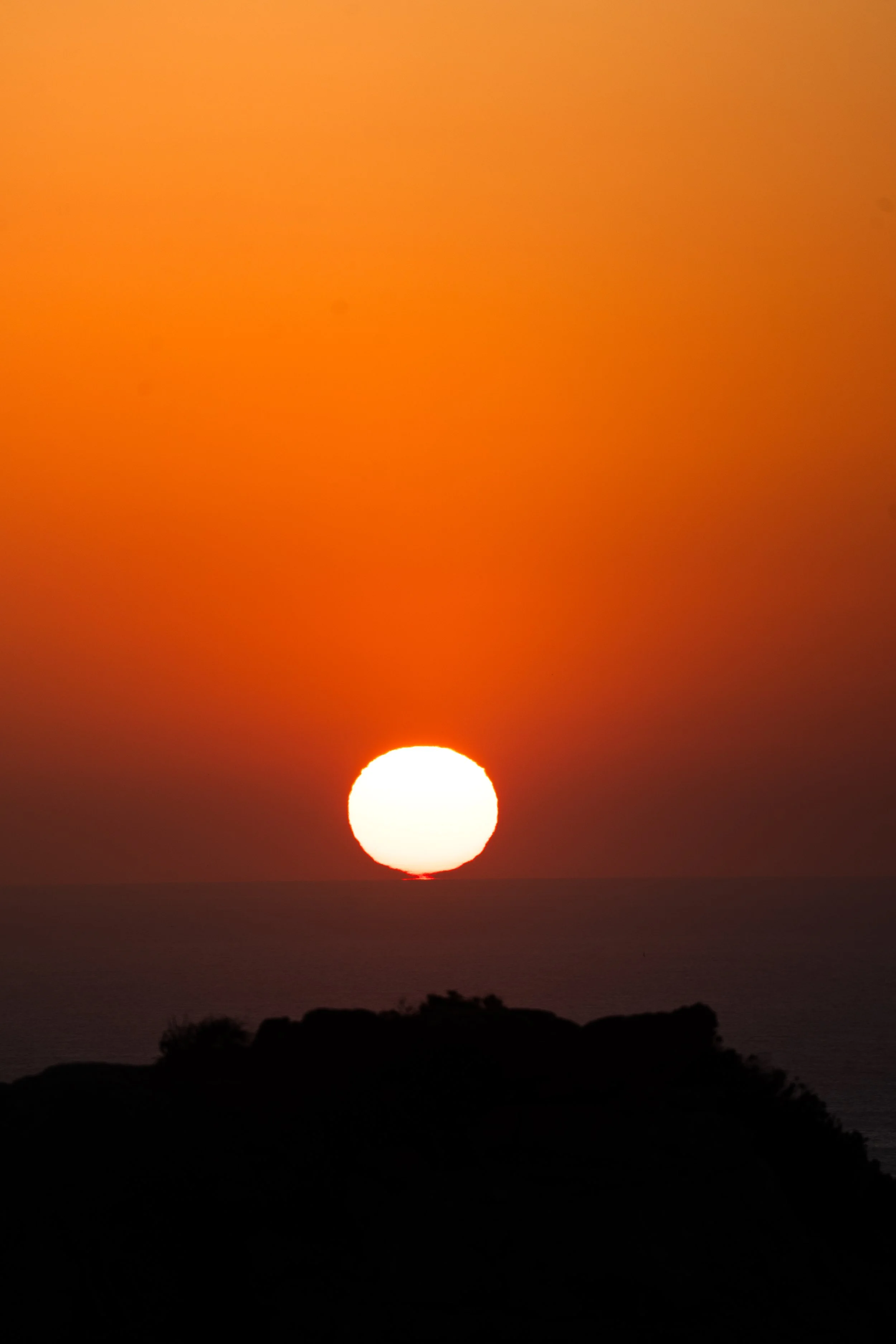 Coucher de soleil sur l'océan avec un ciel orange et un soleil blanc à l'horizon, silhouettes de collines en premier plan.