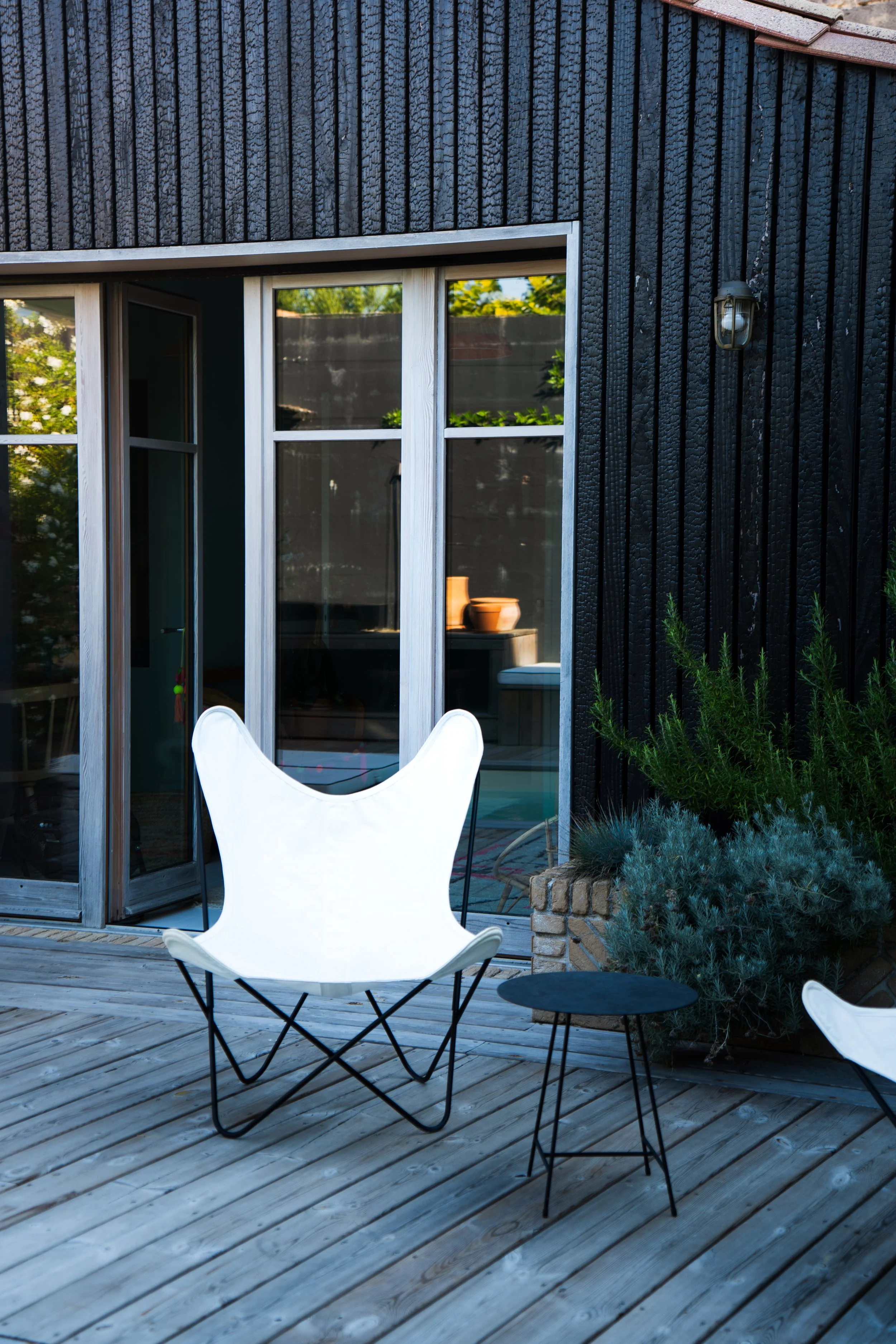 Terrasse en bois avec des meubles en métal noir, un fauteuil blanc en forme d'oiseau, une petite table ronde noire, et un mur extérieur noir avec des fenêtres et des plantes vertes.