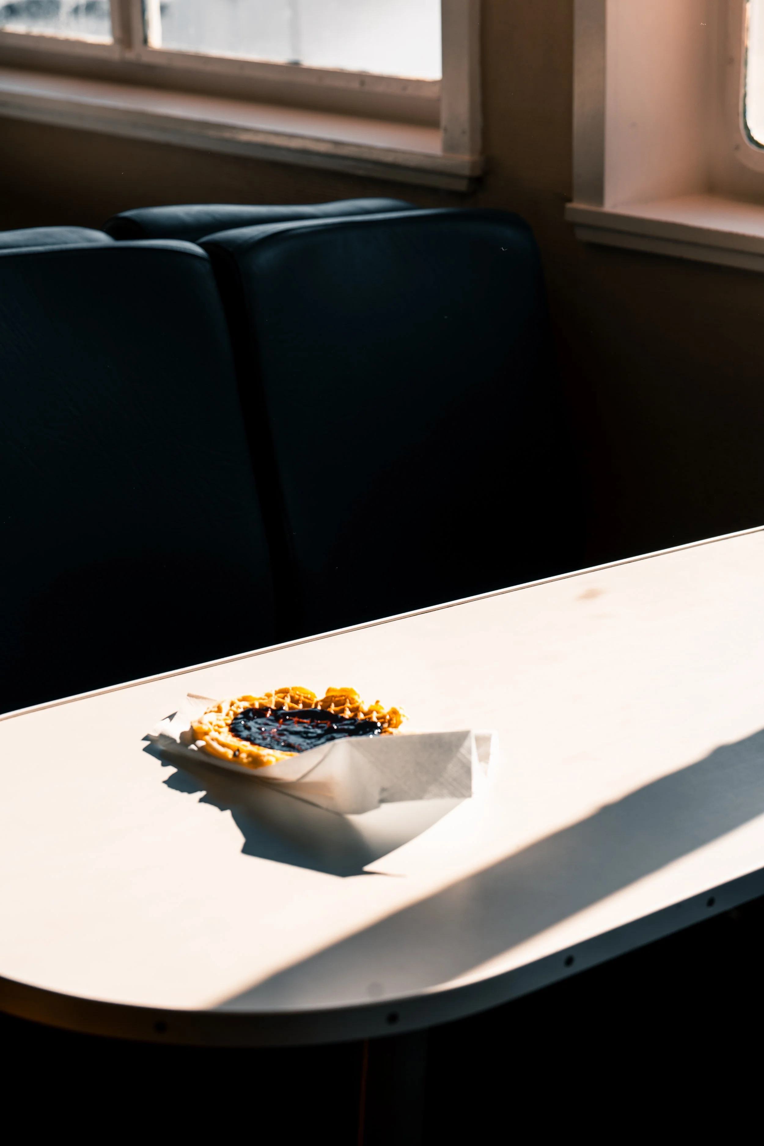 Une tartelette aux fruits, apparemment aux myrtilles, sur une assiette en papier sur une table blanche, avec une fenêtre en arrière-plan laissant entrer la lumière