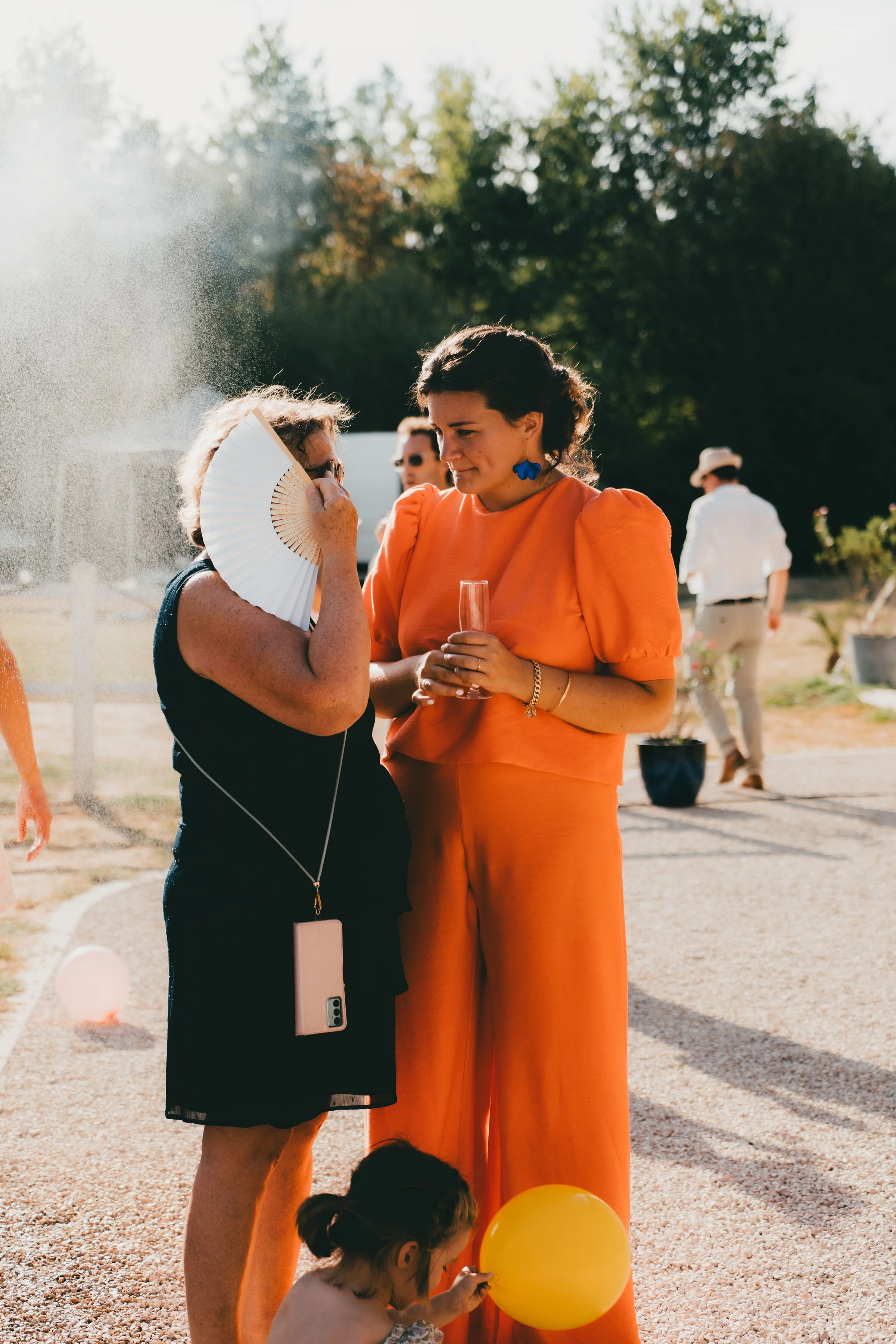 Deux femmes discutent lors d'une fête en plein air, une d'elles porte une robe orange avec des boucles d'oreilles bleues, l'autre une robe noire et tient un éventail blanc. Une fillette avec une balle jaune est assise à leurs pieds. En arrière-plan, d'autres personnes et des arbres sont visibles.