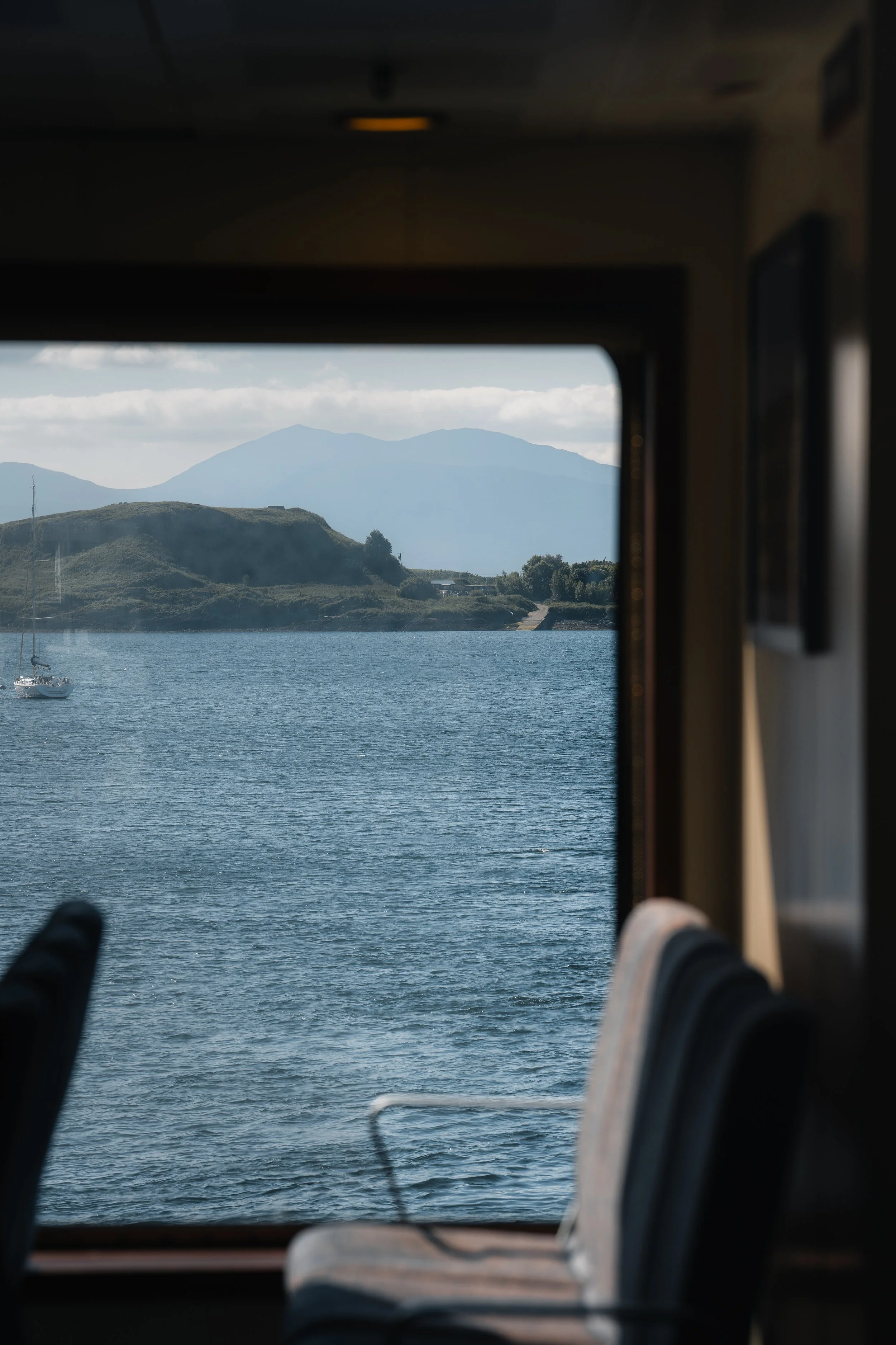 Vue depuis l'intérieur d'un bateau montrant un paysage avec de l'eau, des collines et des montagnes au loin, à travers une fenêtre.
