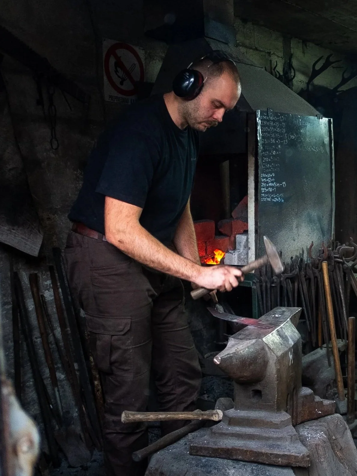 Man black t-shirt, brown pants, and ear protection working at an anvil with a hammer in a blacksmith shop, with tools and a chalkboard in the background.