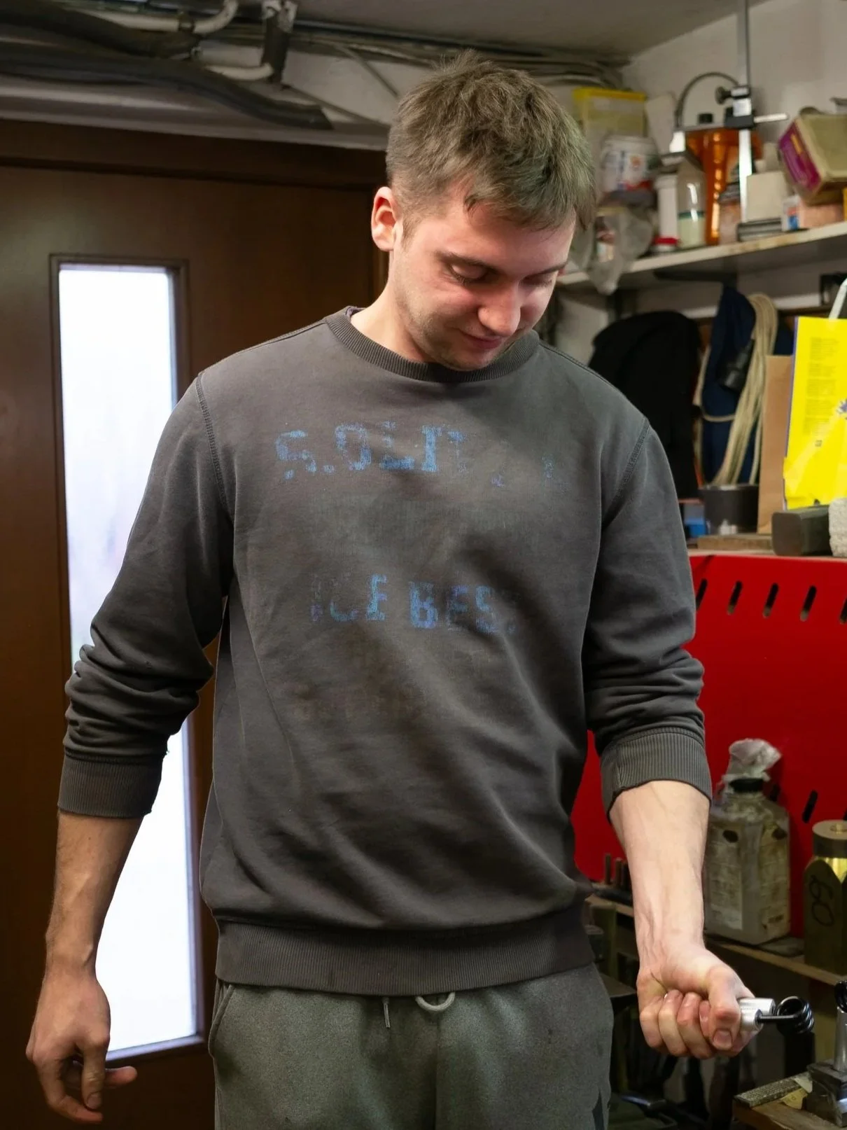 A young man with short brown hair, wearing a dark gray sweatshirt and gray sweatpants, holding a metal tool in his right hand in a workshop with shelves and tools in the background.