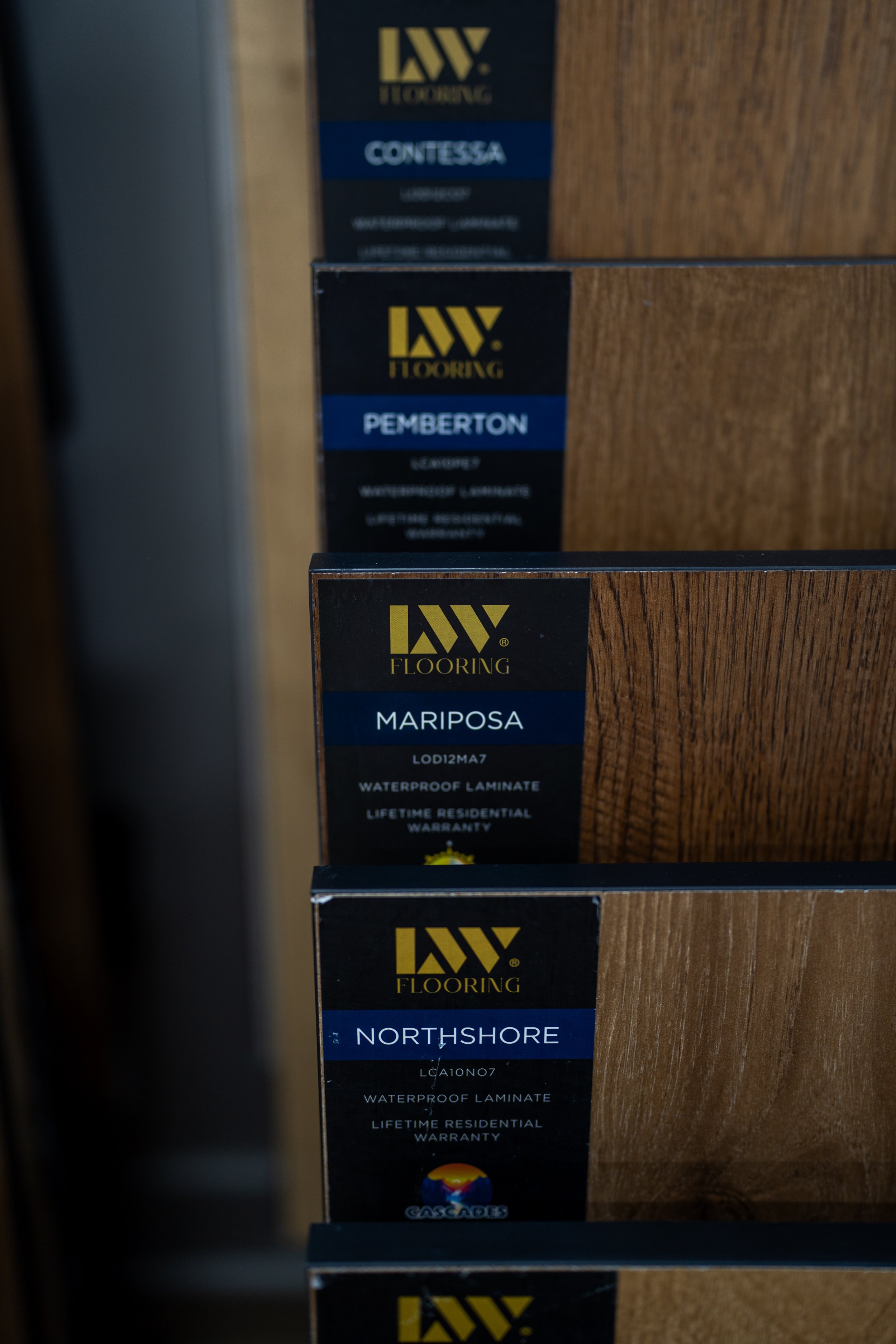 Display of flooring samples with labeled wood textures and names, including 'Mariposa', 'Northshore', and 'Pemberton', on black tags with yellow and white text.