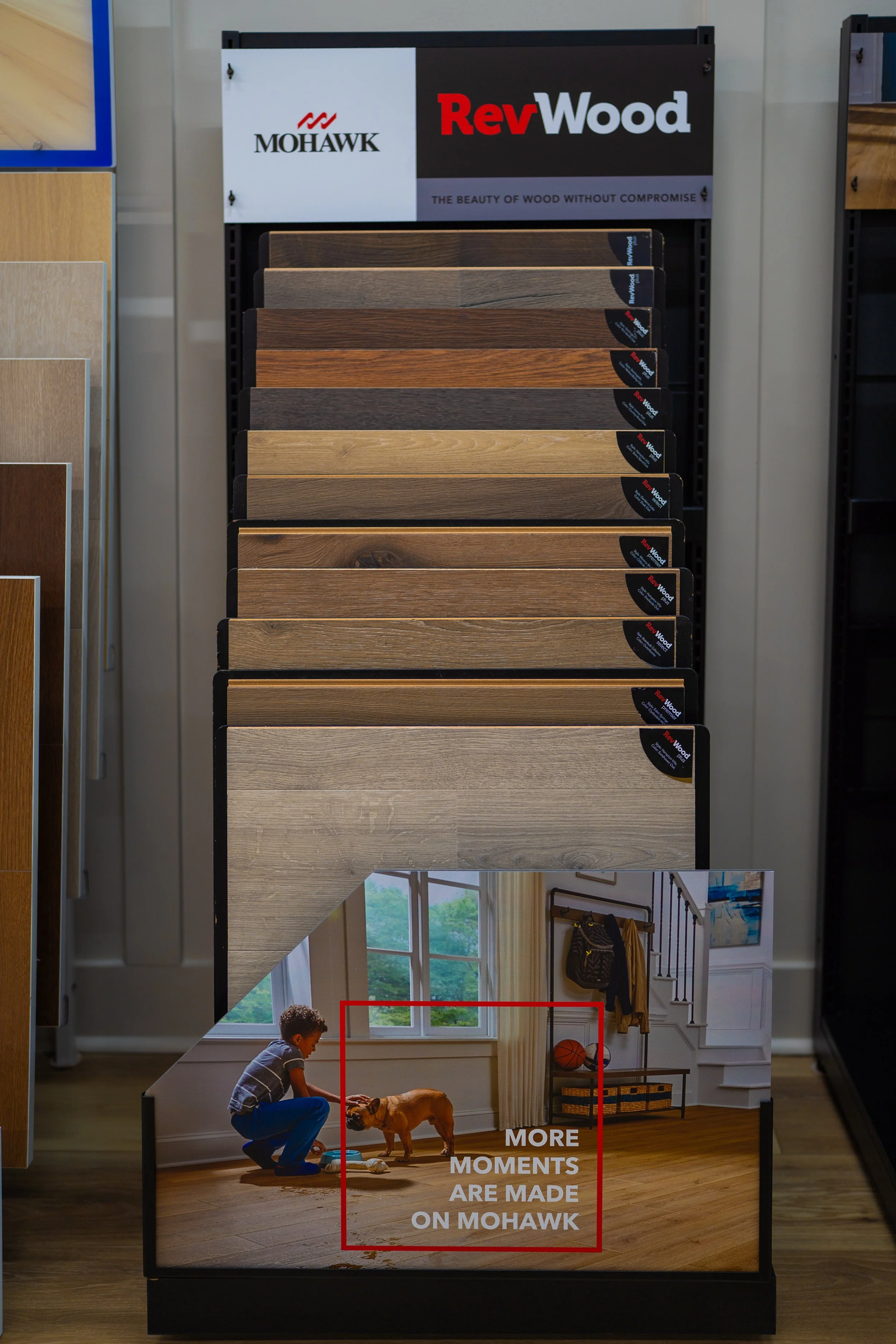 Display of Mohawk hardwood samples with a sign that reads 'RevWood' and a tagline 'The beauty of wood without compromise,' featuring an image of a child and dog in a living room with the words 'More moments are made on Mohawk'.