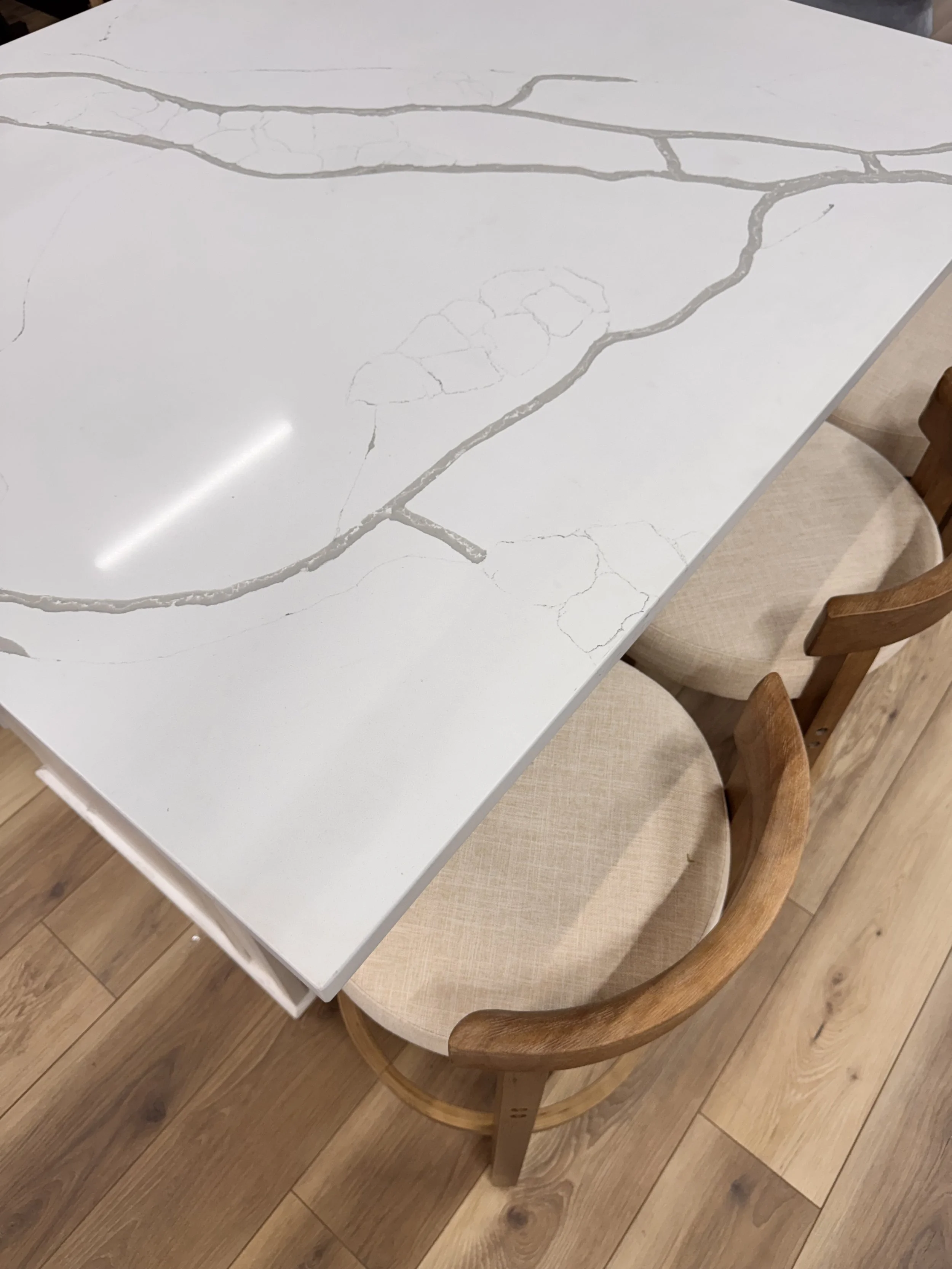 Close-up of a white marble table with gray veining, surrounded by wooden chairs with beige upholstered seats, on a wooden floor at Seamless Floors showroom located in St Augustine.