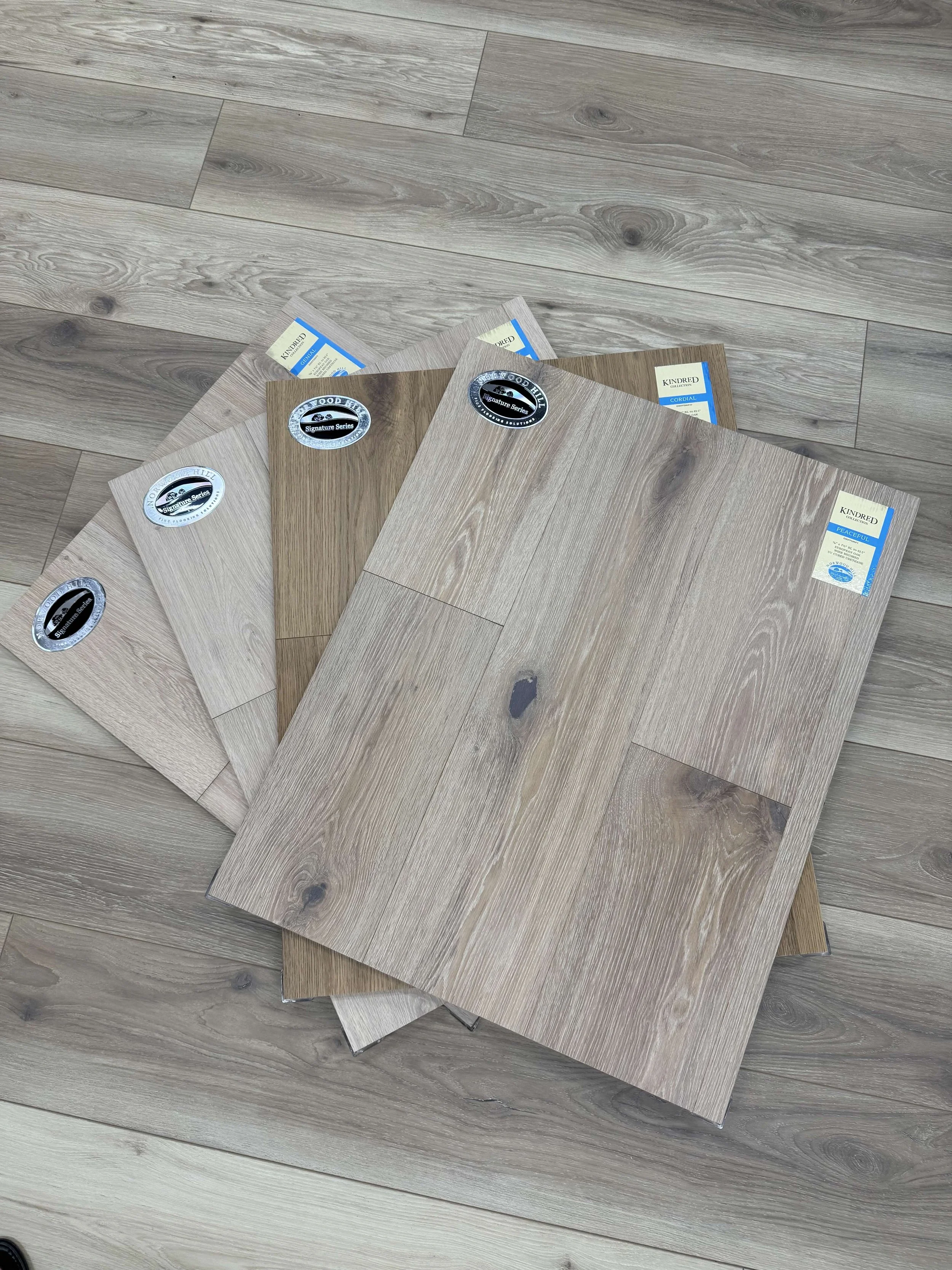 Four wood laminate flooring samples in different shades of brown and beige, fanned out on a light wood floor. Each sample has a label and a black and silver round sticker.
