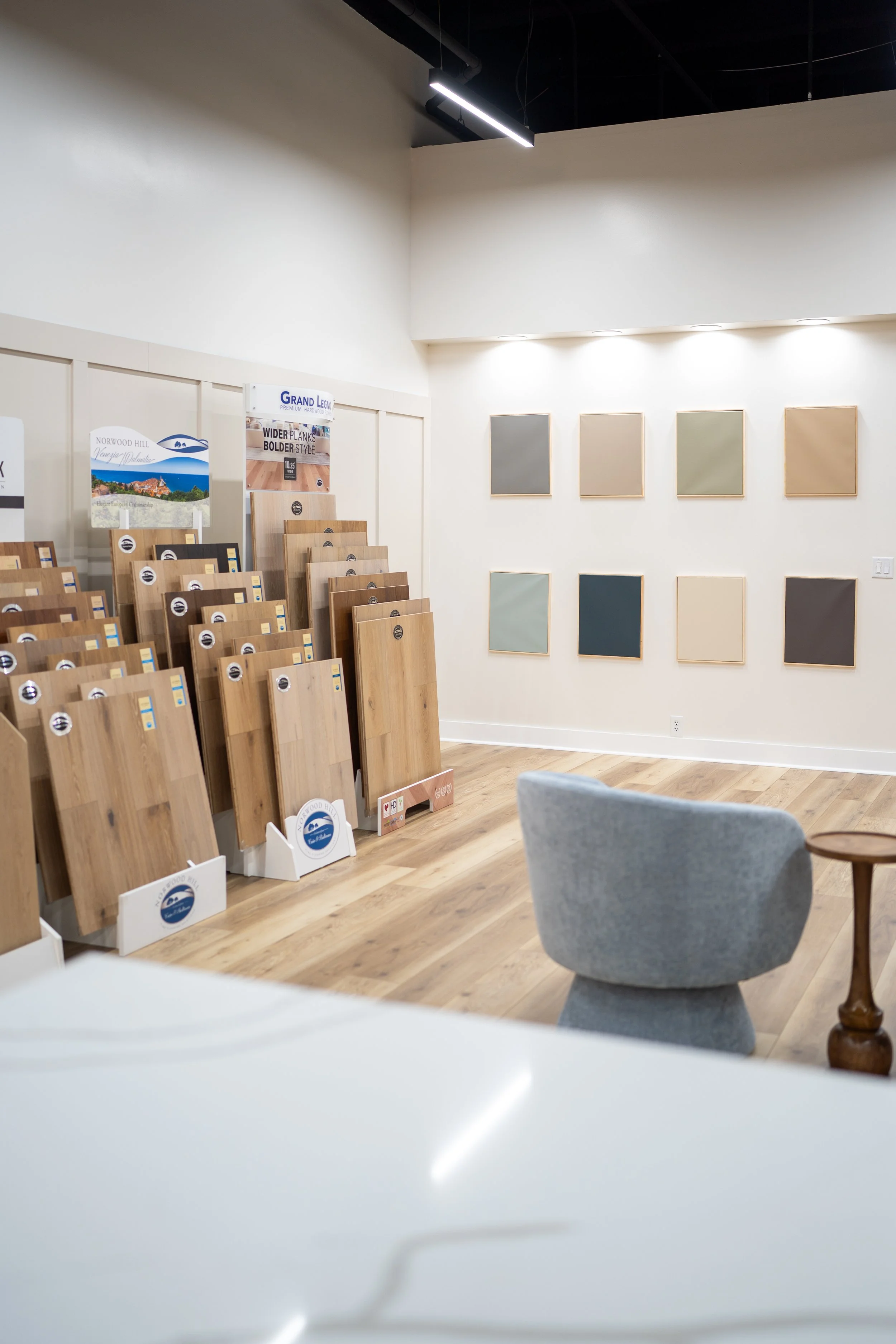 Showroom with display of hardwood flooring samples on the wall and stands of flooring samples on the left side, with a gray armchair and small wooden side table in the foreground.
