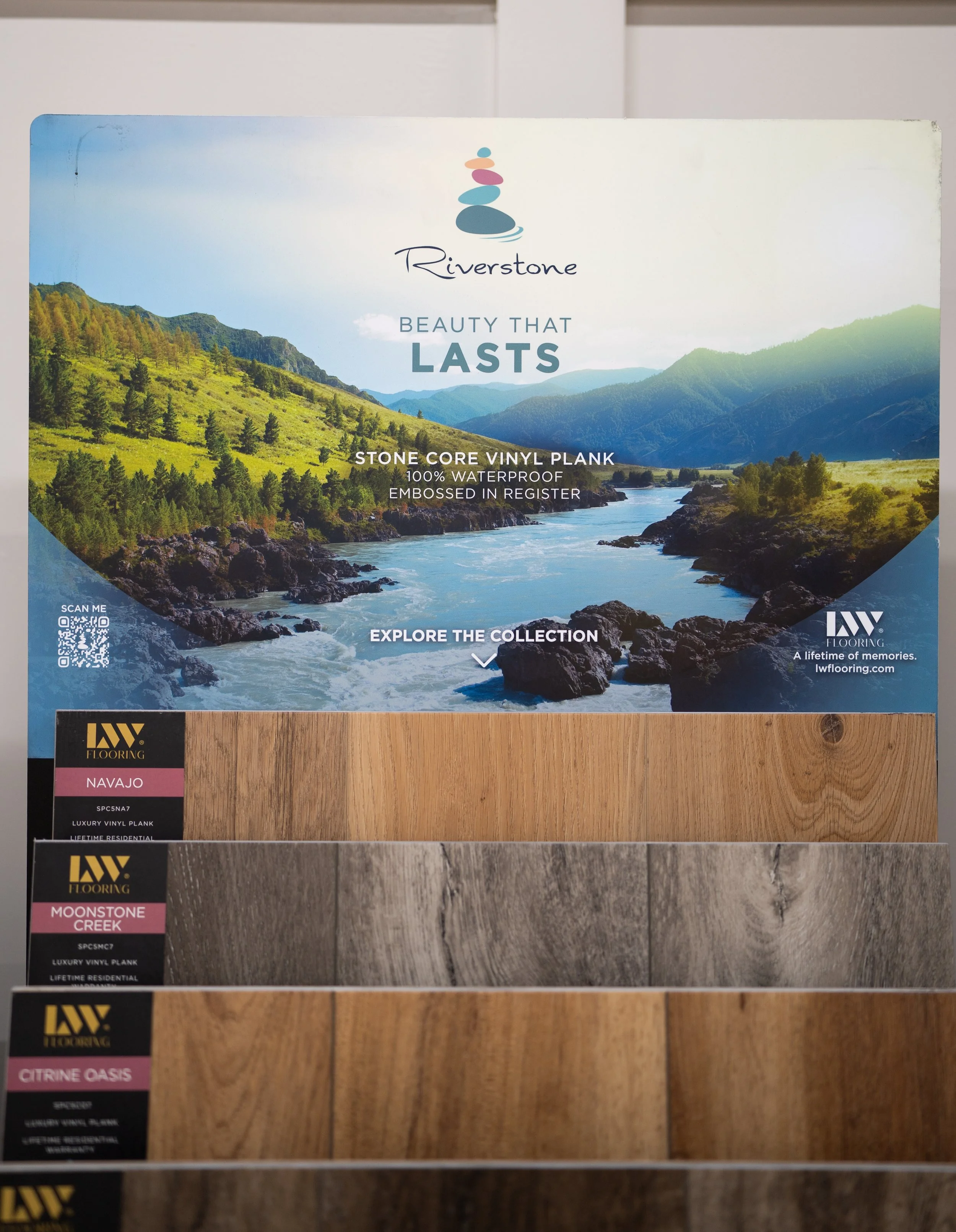 Display of flooring samples with a scenic Riverstone advertisement on top, showcasing a mountain river landscape and wood plank samples labeled Navajo, Moonstone Creek, and Citrine Oasis.