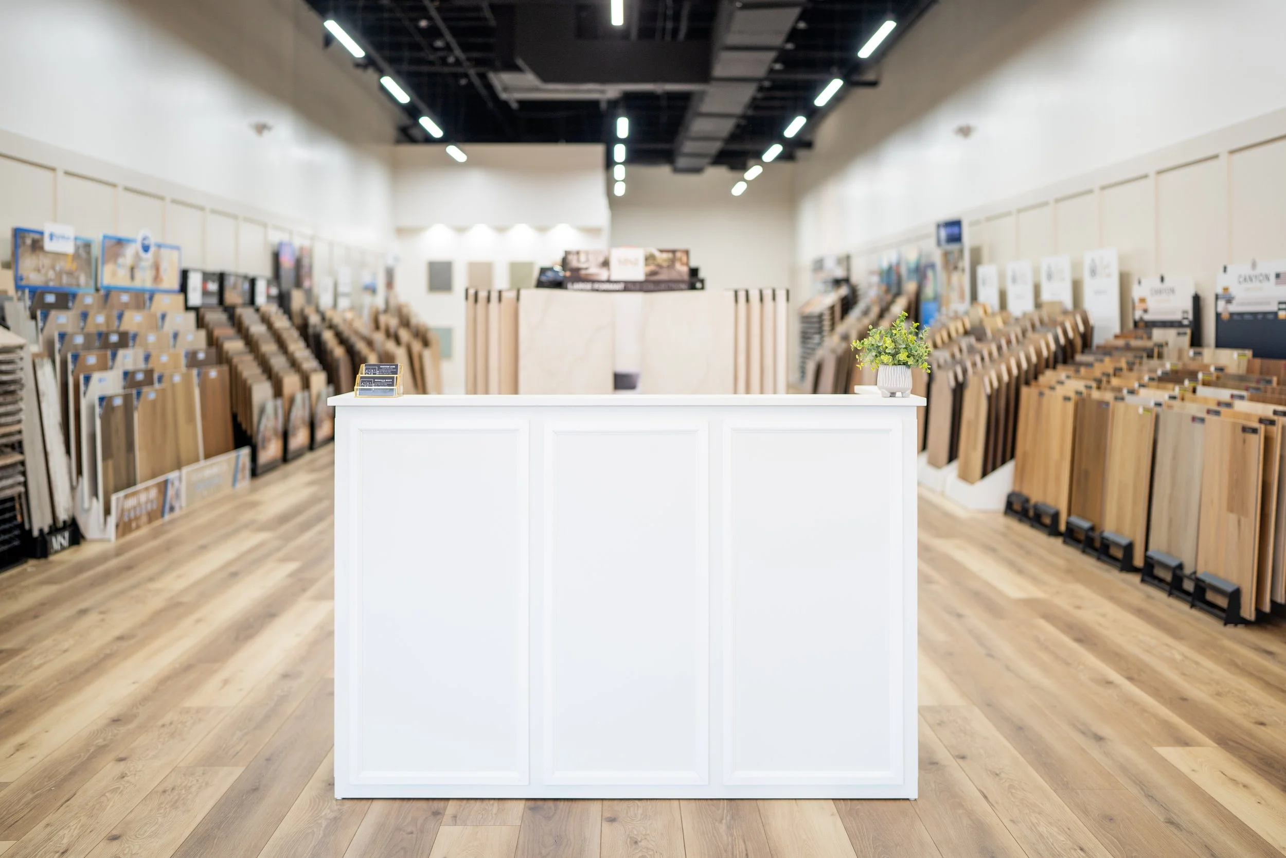 Interior of a flooring store showcasing various wood flooring samples on display racks along the walls. A white counter with a small potted plant is in the foreground.
