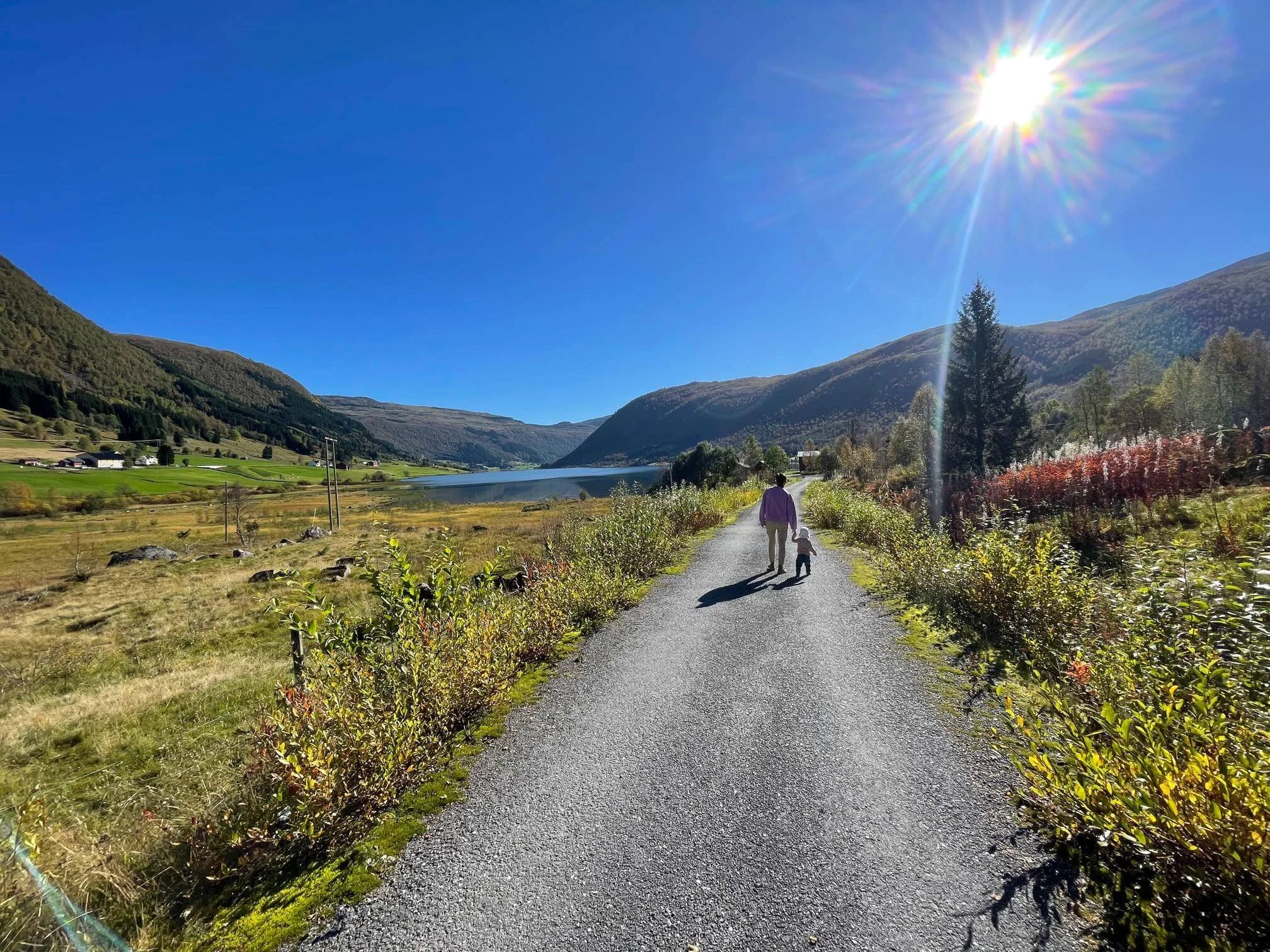 Une mère et un enfant marchent sur un chemin au bord d'un lac entouré de montagnes sous un ciel clair et ensoleillé.