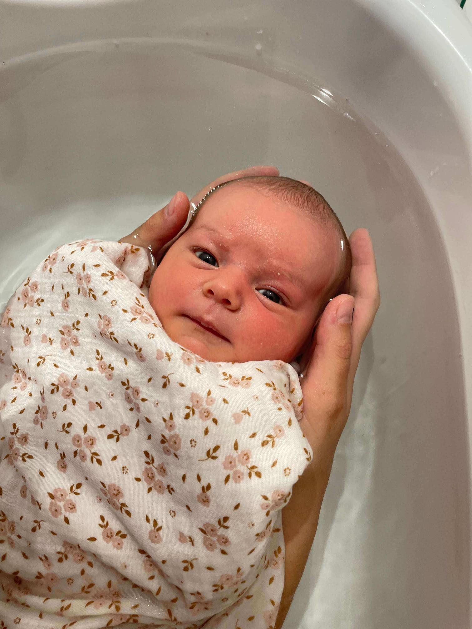 Un bébé est tenu dans une main, enveloppé dans une couverture à motifs floraux roses et marrons, en train de prendre un bain dans une baignoire blanche.