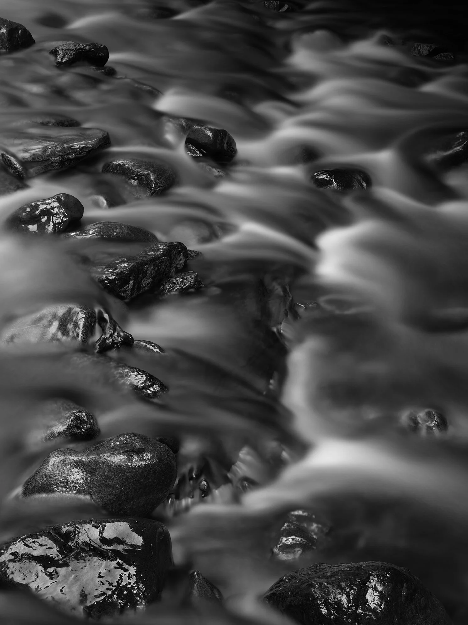 Black and white photo of water flowing over black rocks on a riverbed with long exposure effect creating a smooth appearance.