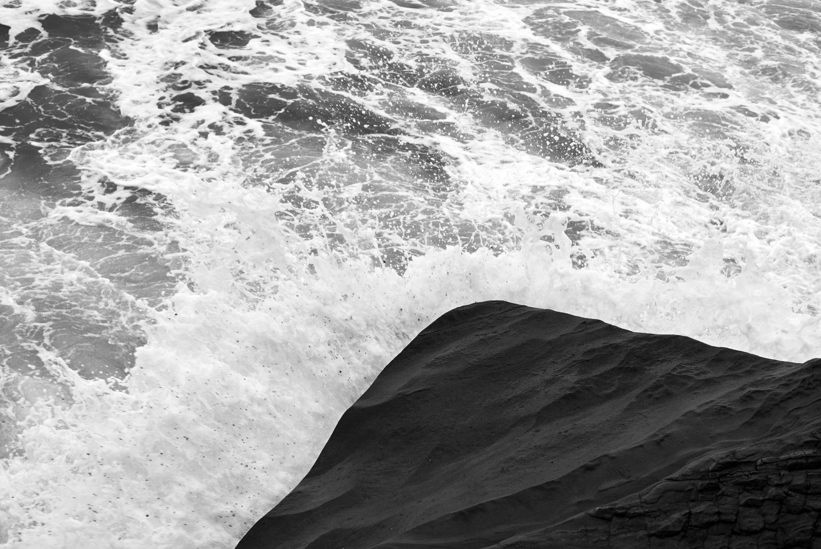 A black rock on the ocean with white foamy waves crashing against it.