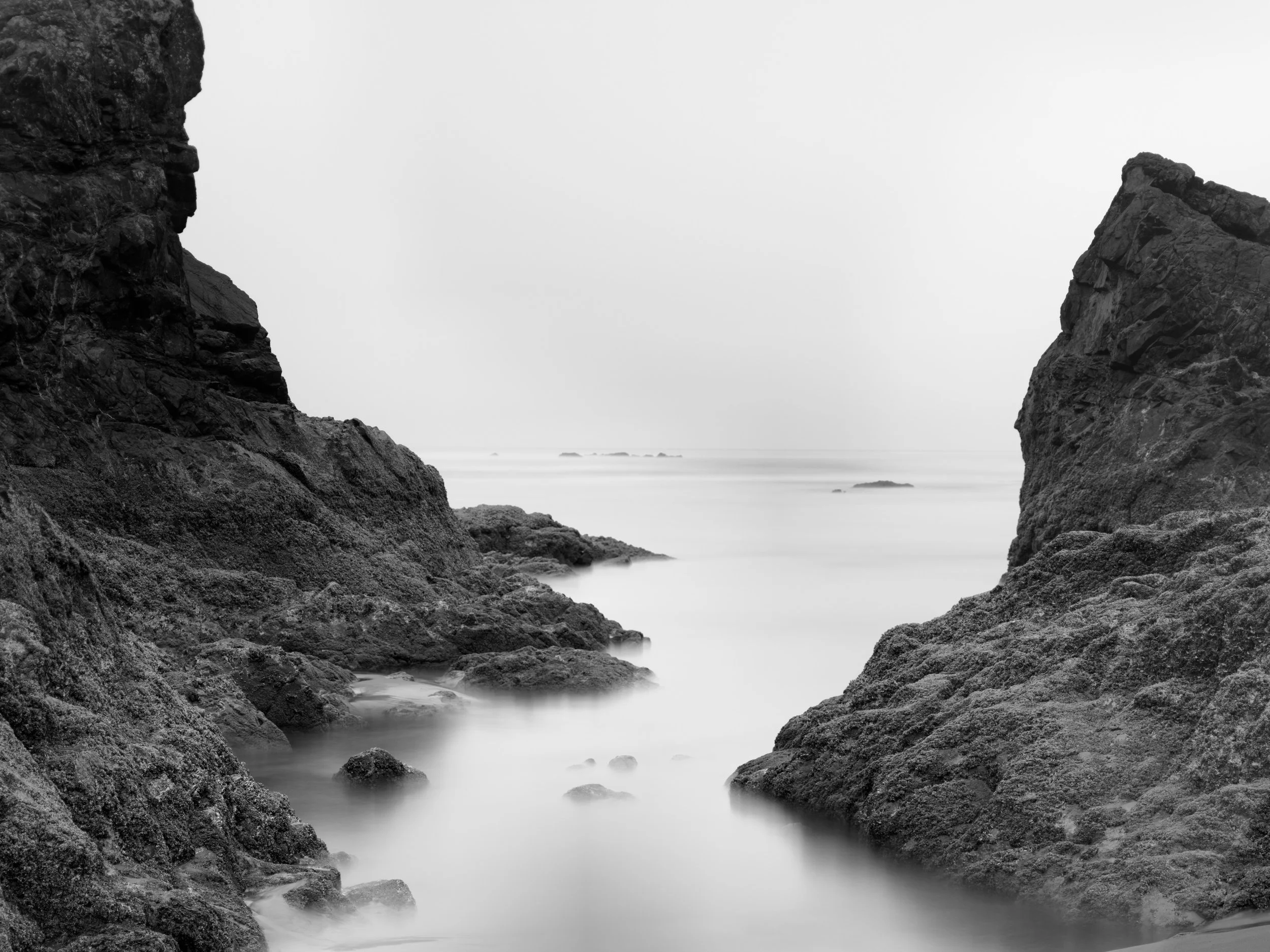 Black and white photograph of a rugged coastal scene with rocky cliffs on either side and water flowing between them, creating a misty effect.