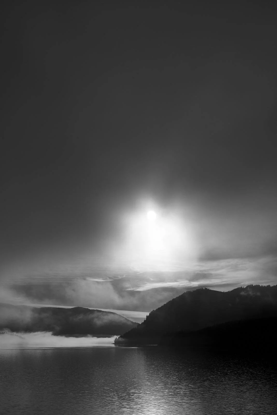 Black and white photo of a scenic lake with surrounded hills and mist, under a bright sun in the sky.