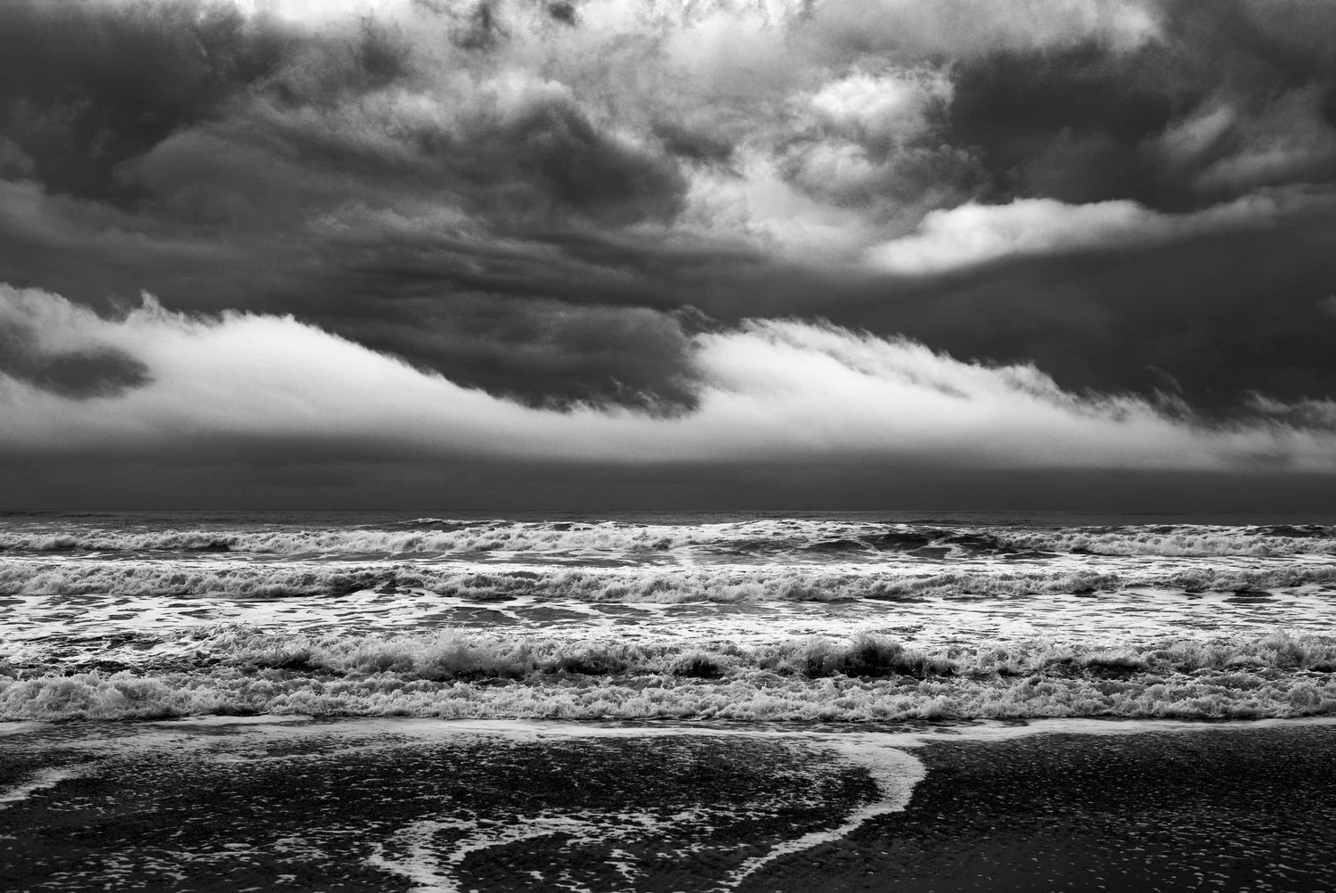 Black and white photo of a stormy beach with dark clouds overhead and waves crashing on the shore.