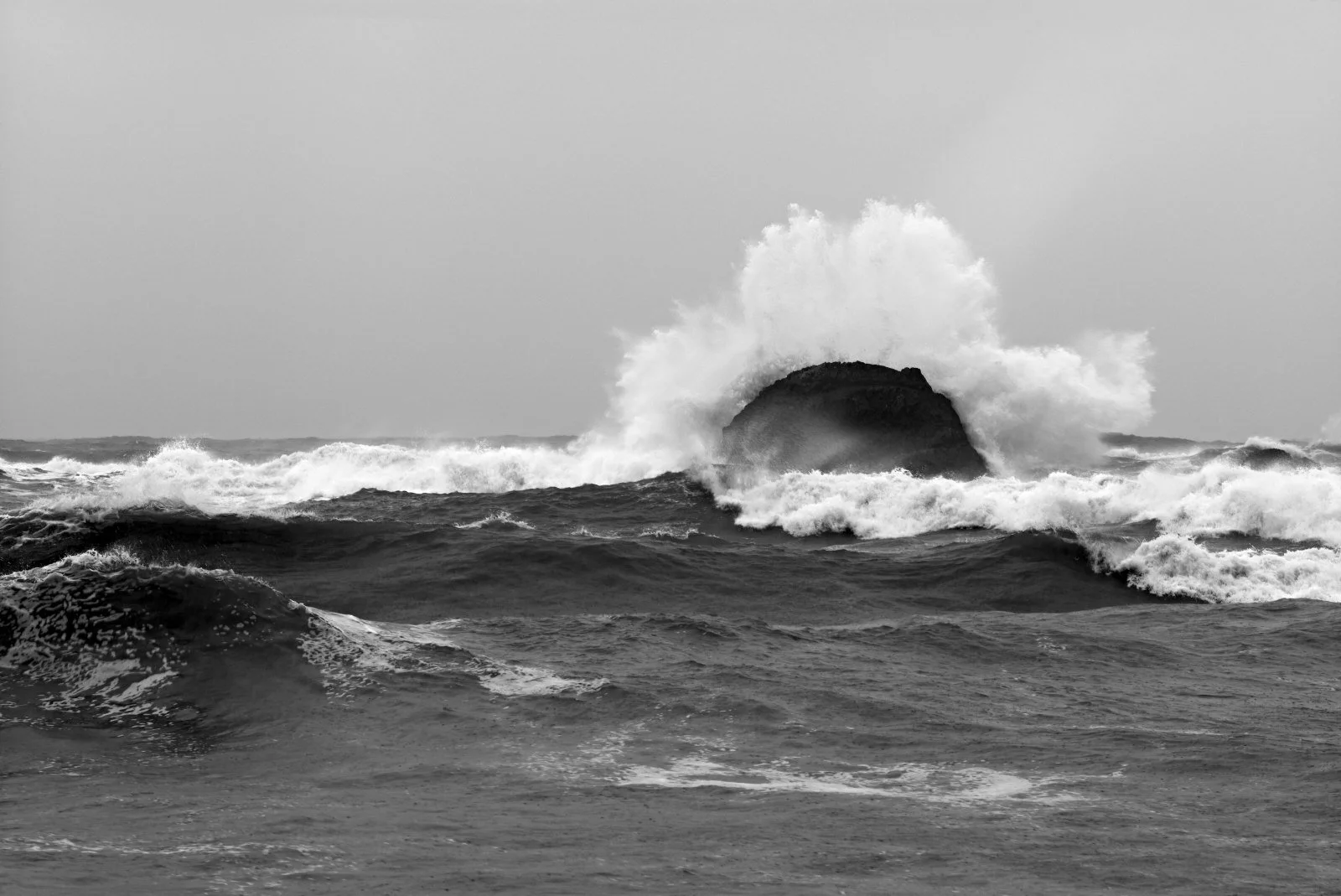A large ocean wave crashing against a rock, with foam splashing into the air, in black and white.