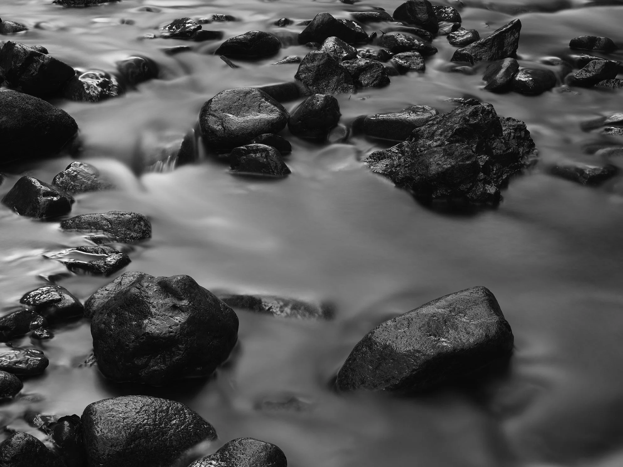 Black and white photo of rocks in water with a long exposure effect, creating a smooth motion of the flowing water around the rocks.