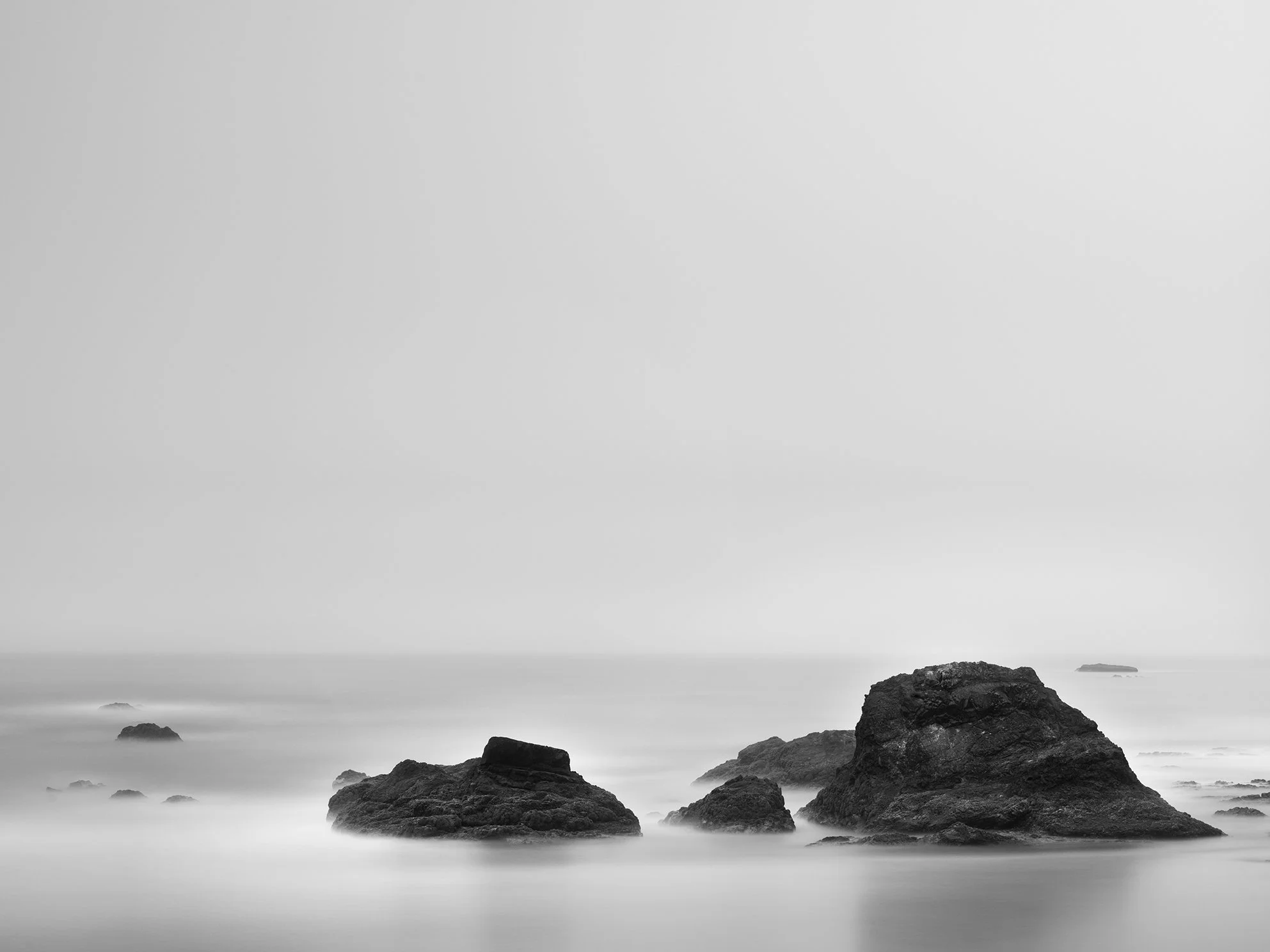 Black and white photo of rocky shoreline with large rocks in the foreground and ocean waves in the background.